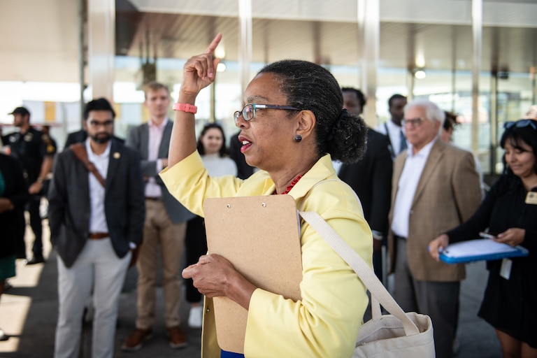 State delegate Robbyn Lewis, wearing a yellow jacket and raising her right hand, addresses a small crowd of people.