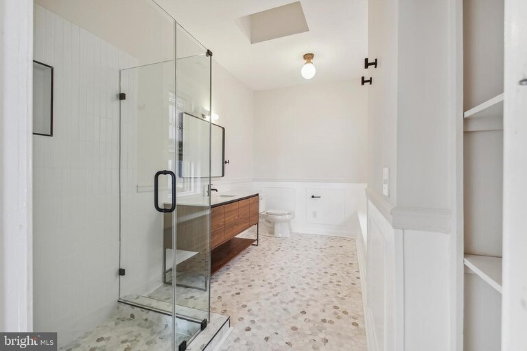 A photo of a large bathroom with a glass-walled shower, light gray walls, white wainscoting includes modern sink fixtures and a modern light fixture.