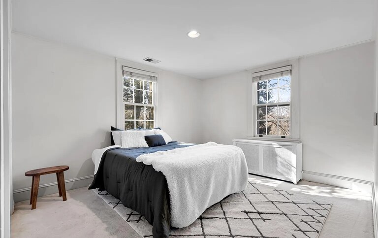 A sparsely decorated bedroom with white walls a white area rug, a small wooden table and a bed that takes up most of the room decorated with a dark gray bed spread.