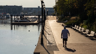 A silhouetted man walks along the harbor with nobody else in sight. Street lights divide the photo in half between the water and the walkway.