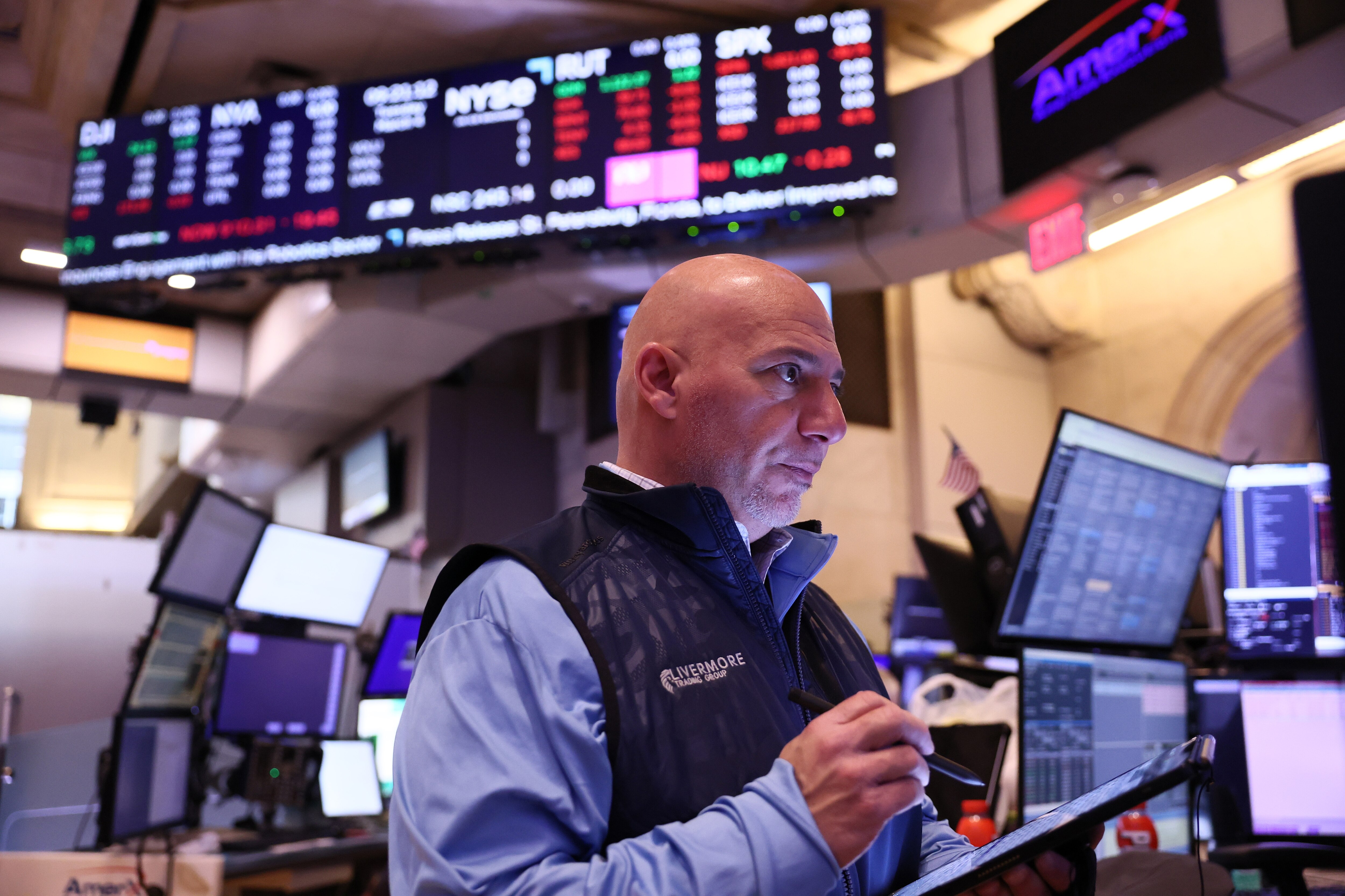 Stock trader working at the New York Stock Exchange. A bald, middle-aged trader wearing a light blue shirt and dark vest with 'Avermore' logo stands in front of multiple computer screens and a large stock market display board showing red and green stock price tickers.