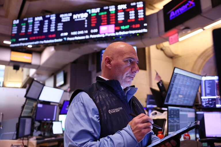Stock trader working at the New York Stock Exchange. A bald, middle-aged trader wearing a light blue shirt and dark vest with 'Avermore' logo stands in front of multiple computer screens and a large stock market display board showing red and green stock price tickers.
