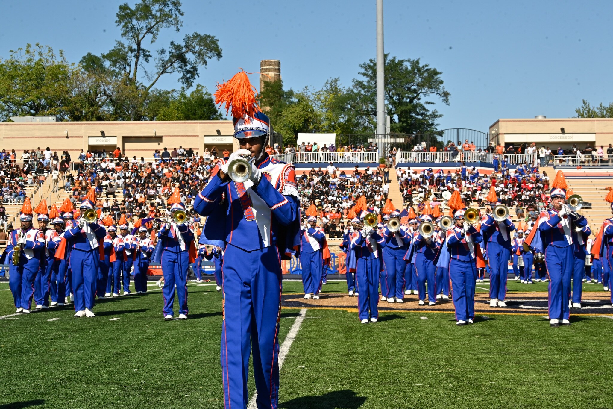 Someone wearing a blue and orange marching band uniform plays a trumpet on a football field in front of the rest of the marching band.