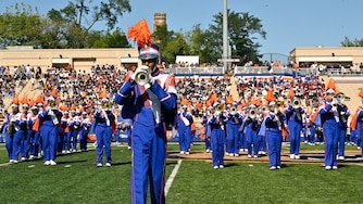 Someone wearing a blue and orange marching band uniform plays a trumpet on a football field in front of the rest of the marching band.