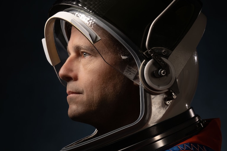 A middle-aged caucasian man wears an astronaut helmet with a visor lifted up and looks off to the left in a portrait against a black background.