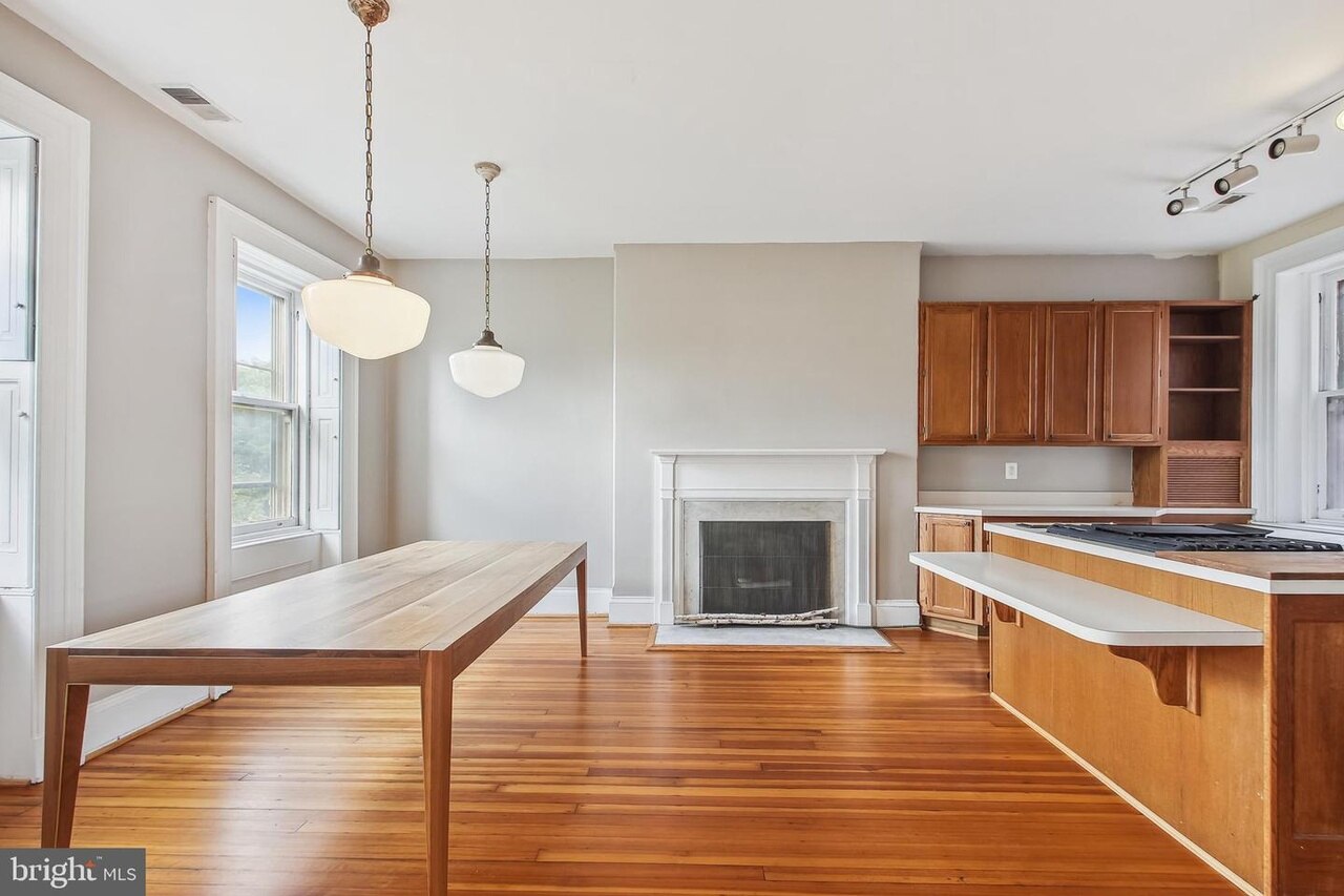A picture of an open concept dining room and kitchen with a large wooden table, pendant light fixtures, a fireplace, and a kitchen counter and cabinets.