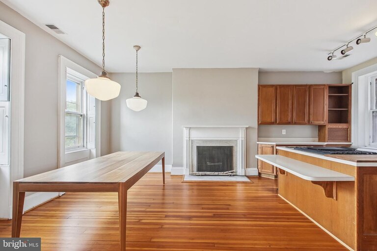 A picture of an open concept dining room and kitchen with a large wooden table, pendant light fixtures, a fireplace, and a kitchen counter and cabinets.