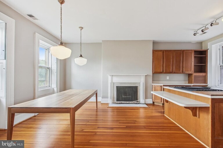 A picture of an open concept dining room and kitchen with a large wooden table, pendant light fixtures, a fireplace, and a kitchen counter and cabinets.