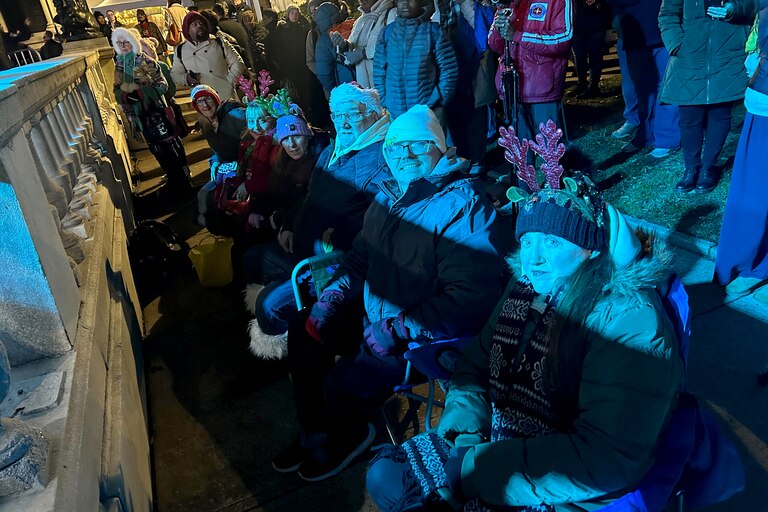 The Milligan family sit in folding chairs in the front row of the stage as festivities start for the 2025 Monument Lighting in Baltimore.