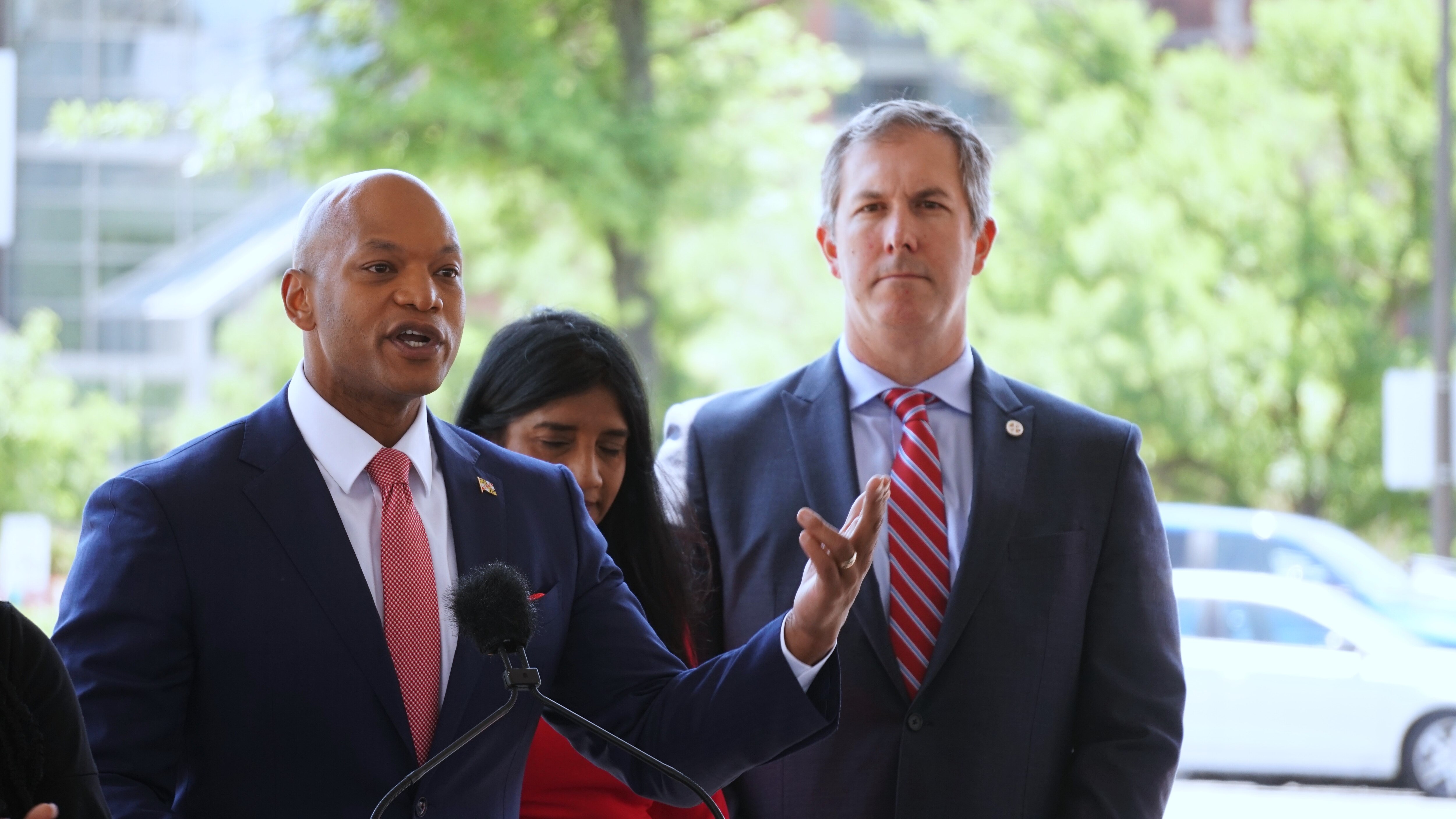 A man in a suit and tie is talking at a podium as a woman and another man in a suit and tie stand behind him.