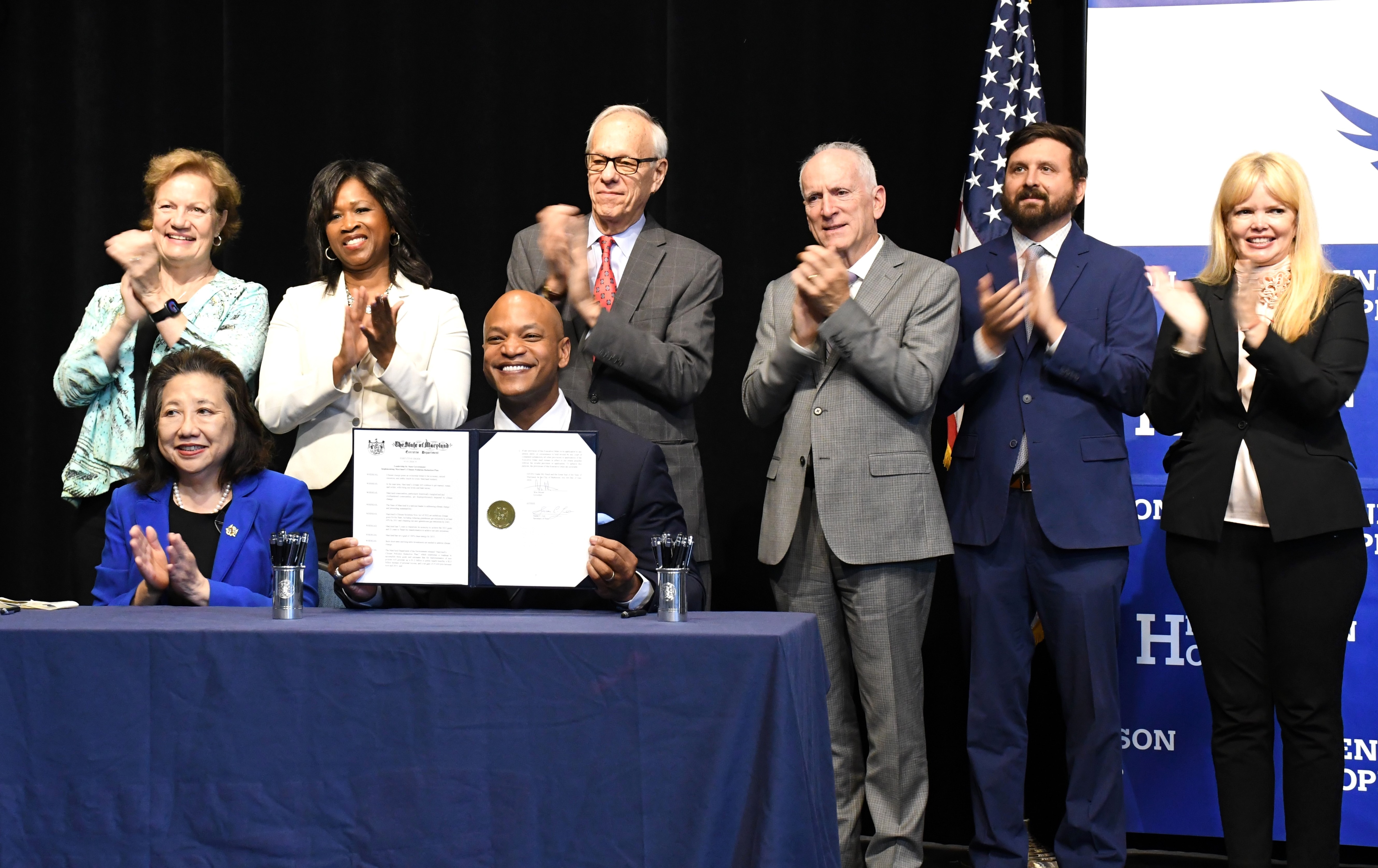 Officials applaud Maryland Gov. Wes Moore after signing an executive order on climate change on Tuesday, June 4, 2024 at Elmer A. Henderson: A Johns Hopkins Partnership School in East Baltimore. Seated next to the governor is Secretary of State Susan Lee. Top row, from left: Kim Coble of the Maryland League of Conservation Voters; Environment Secretary Serena McIlwain; Maryland Energy Administration Director Paul Pinsky; Transportation Secretary Paul Wiedefeld; Natural Resources Secretary Josh Kurtz; and Meghan Conklin, the state's chief sustainability officer.