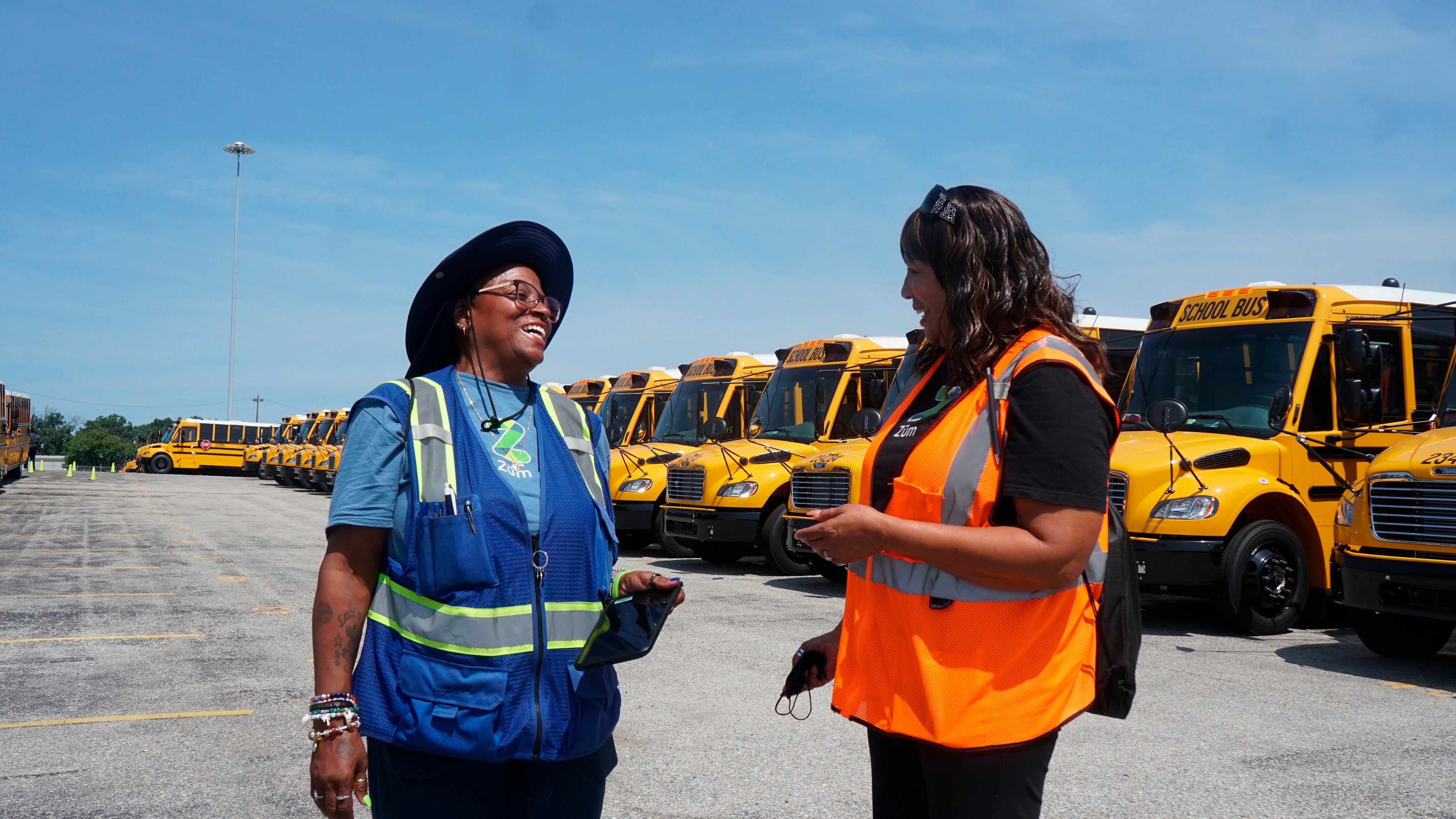 Donna McSee and Jackie Scott, drivers for Howard County Public Schools employed by Zūm, talk in front of a line of brand new yellow school buses at Zūm’s Jessup operations hub on Aug. 22.