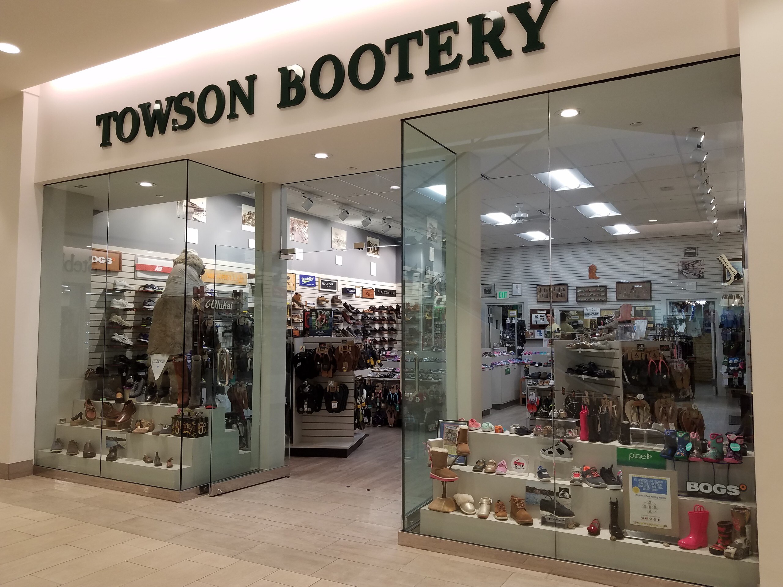 The entrance to the Towson Bootery in The Shops at Kenilworth. Black text says 'Towson Bootery' at the top and below, glass walls with shoes behind them.