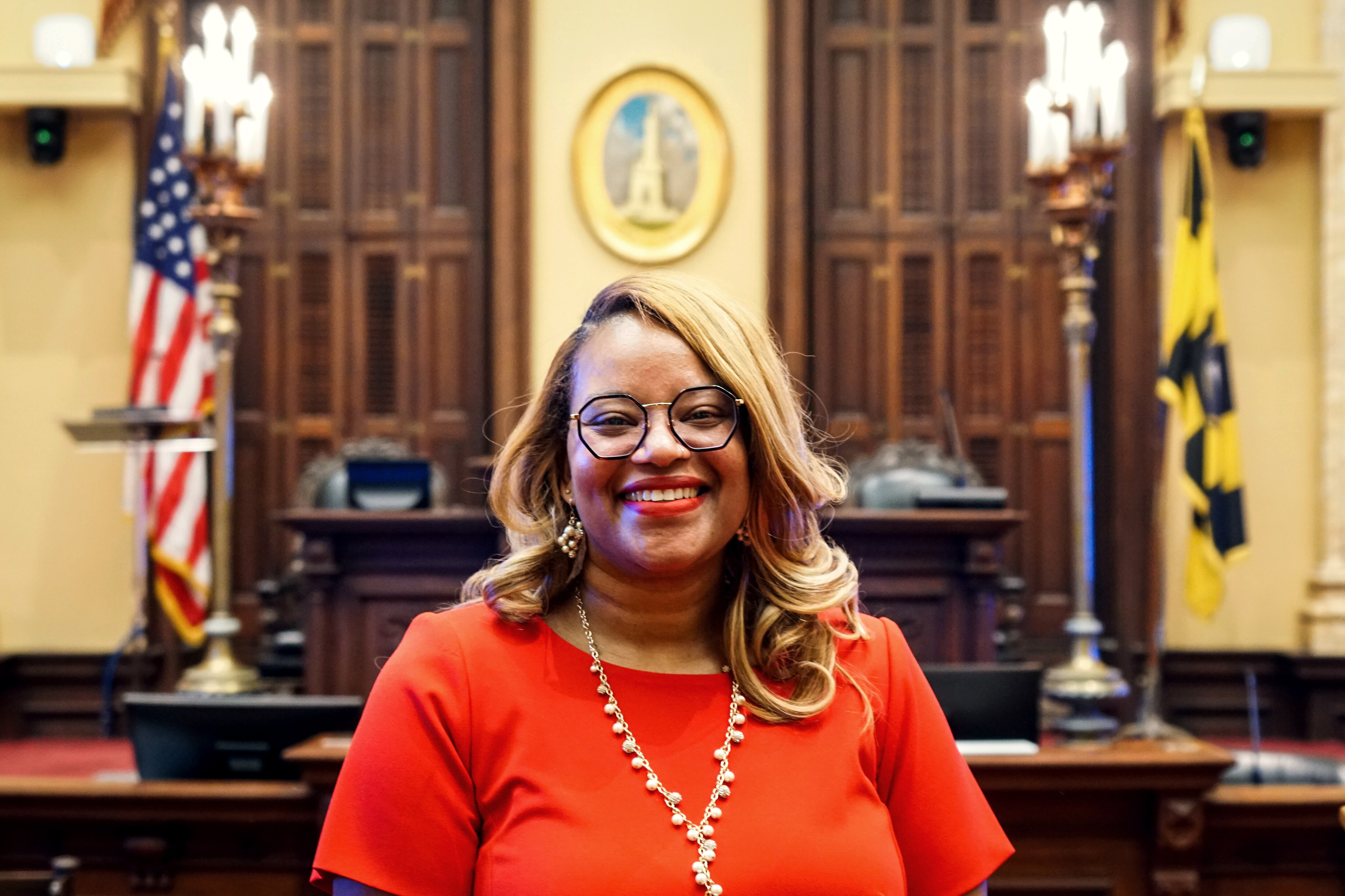 A woman in a red shirt stands in front of a desk and emblem of the City of Baltimore, flanked by a United States flag and a City of Baltimore flag.