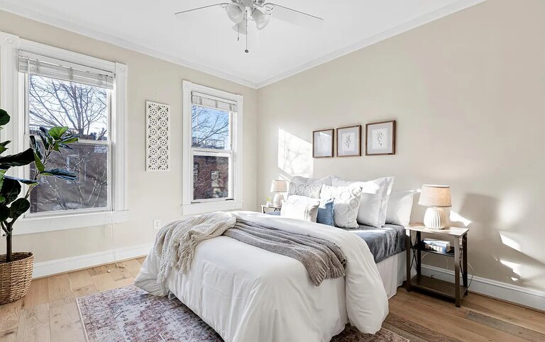 A bedroom with beige walls, two windows and a bed with a white comforter, pillows and blankets.
