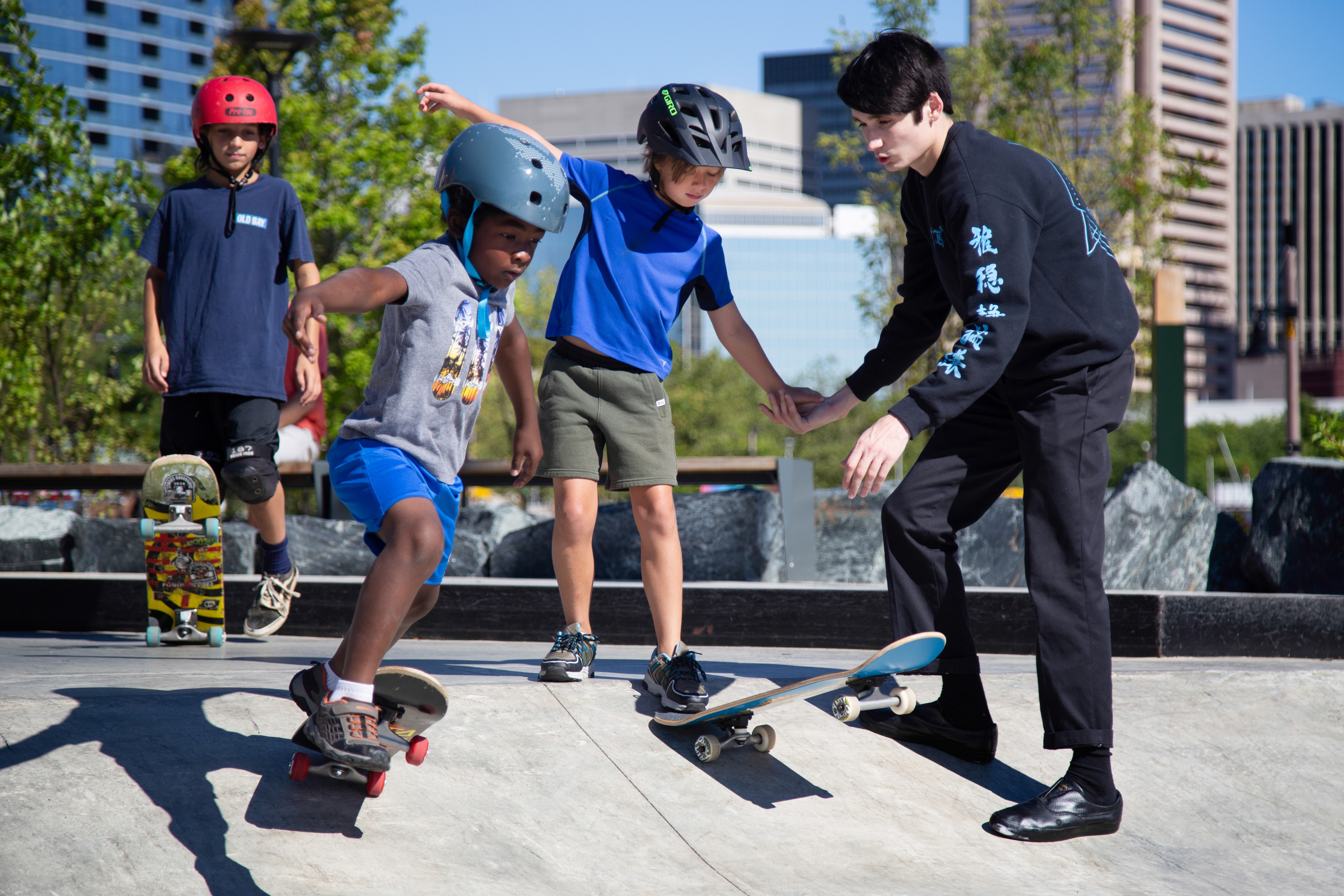 8/13/22 – Baltimore native and professional skateboarder Joey Jett (right) helps teaches Leah Cross, 8, Jeryl Cole, and Hugo Zawaki, 10, how to drop-in on their skateboards. Cross' parents brought her to the park to learn skateboarding from an instructor following numerous driveway crashes from learning on her own. Jett taught the free skateboarding workshop at Rash Field Park on Saturday, August 13th, in collaboration with the Waterfront Partnership of Baltimore.  (Julia Reihs for the Baltimore Banner)