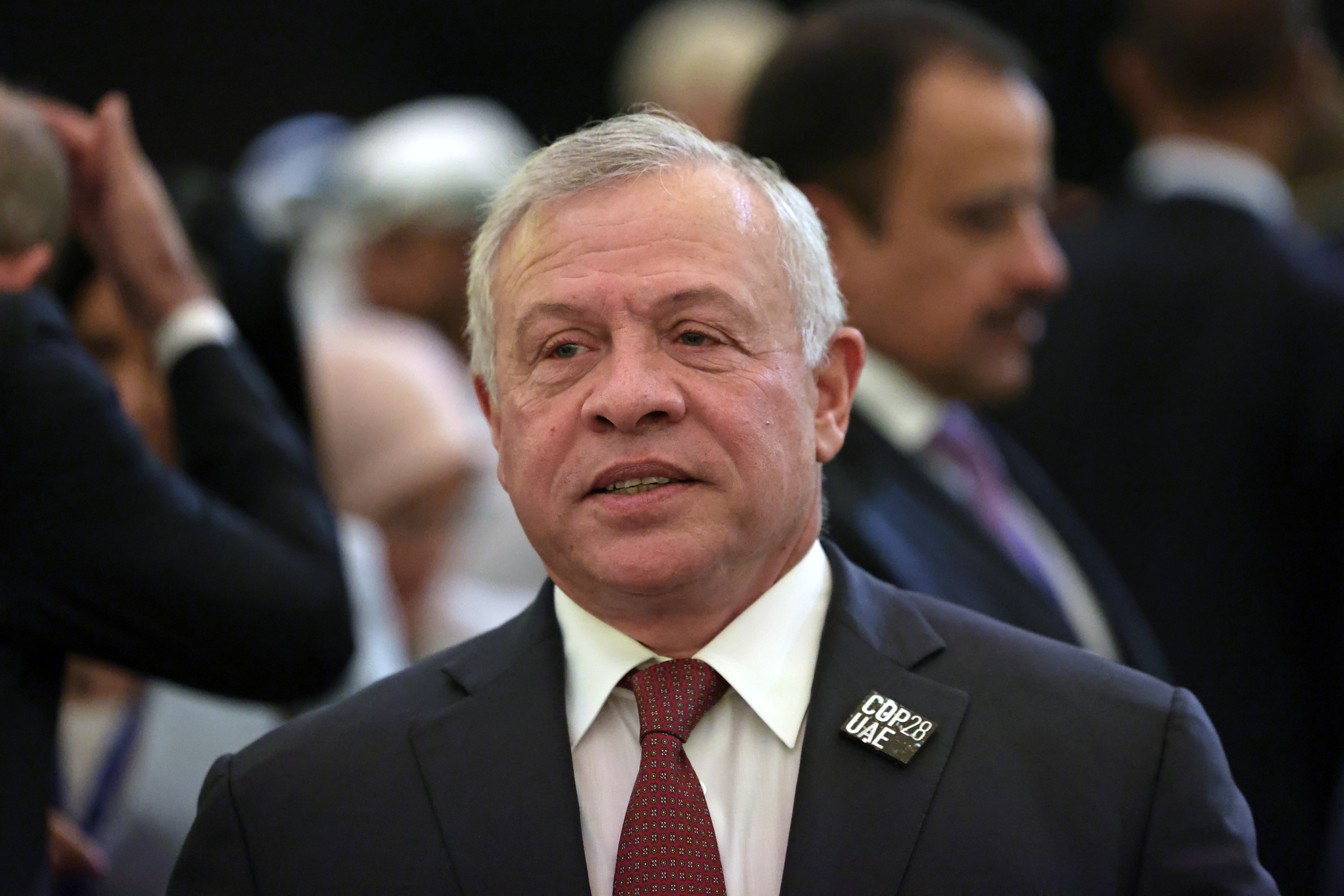 King Abdullah II of Jordan is wearing a dark suit with a dark red tie. He has grey hair and is wearing a lapel pin for a climate summit.