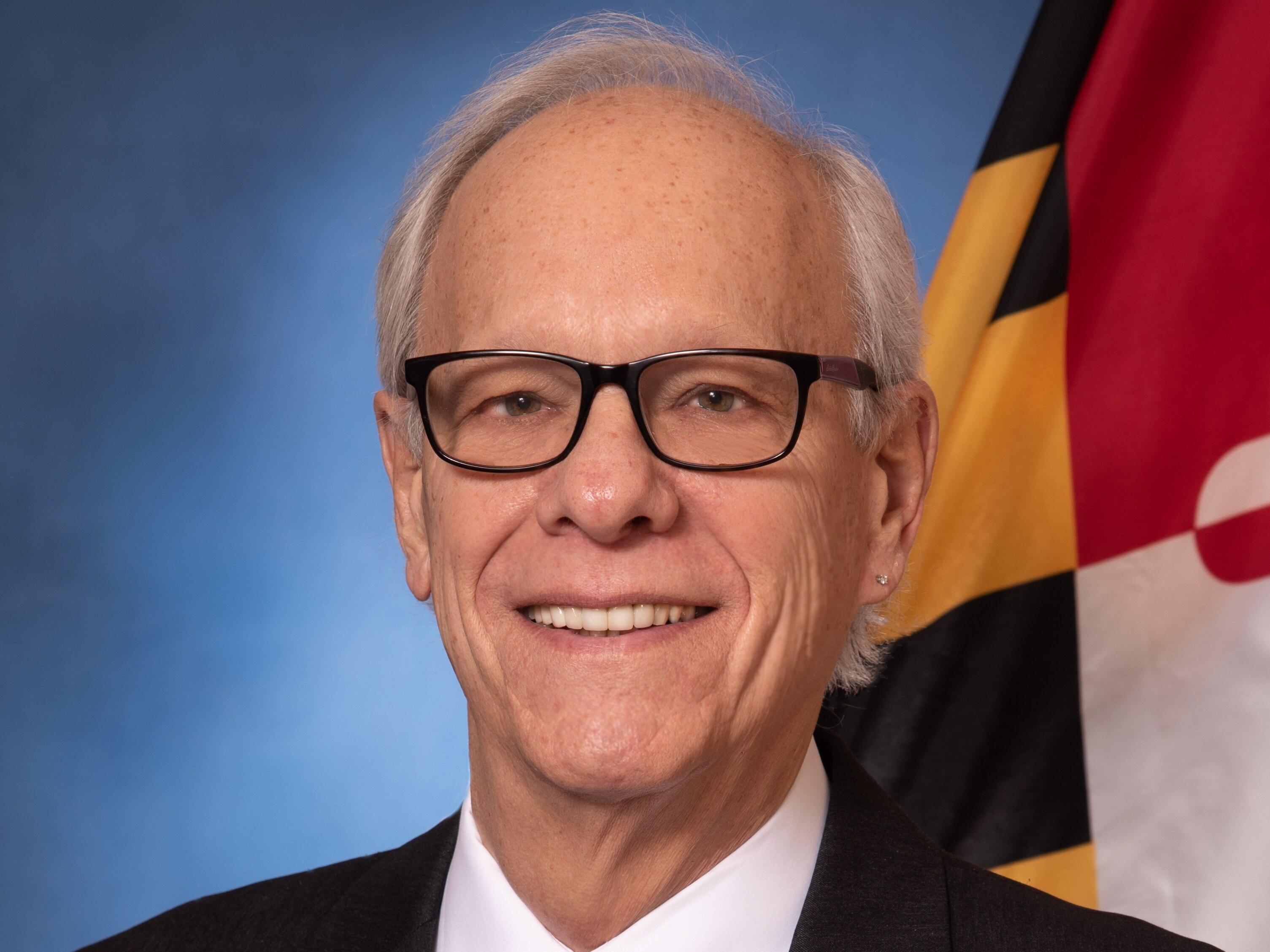 A smiling man with gray hair wearing glasses, a dark suit and a purple tie.