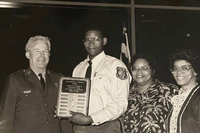 Four people stand posing for a picture as a young man, second from the left, accepts an award.