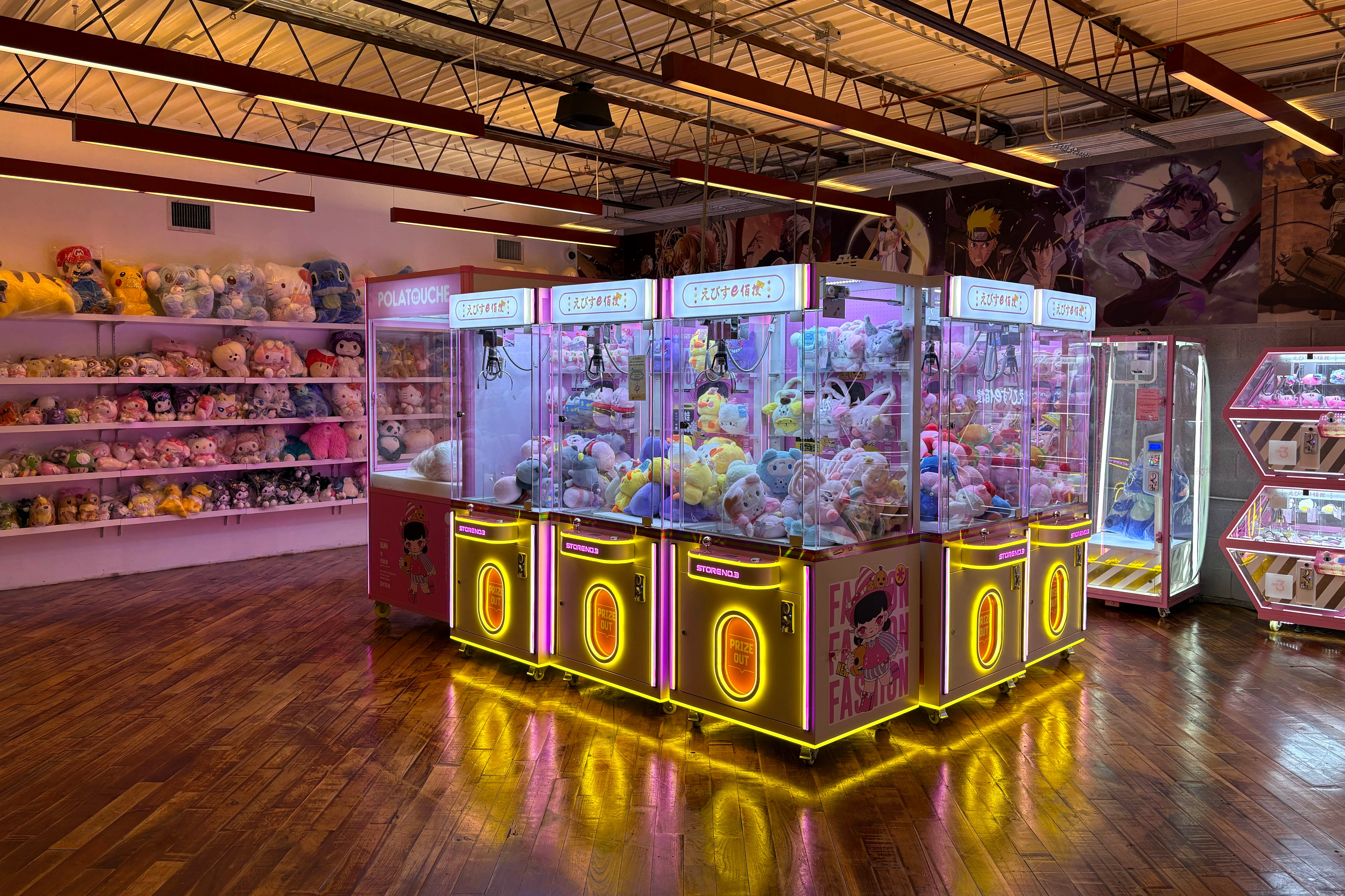 Six claw machines sit back to back in the middle of a large room with wood floors. They are brightly colored and have yellow lights. The back wall features multiple large plush toys.