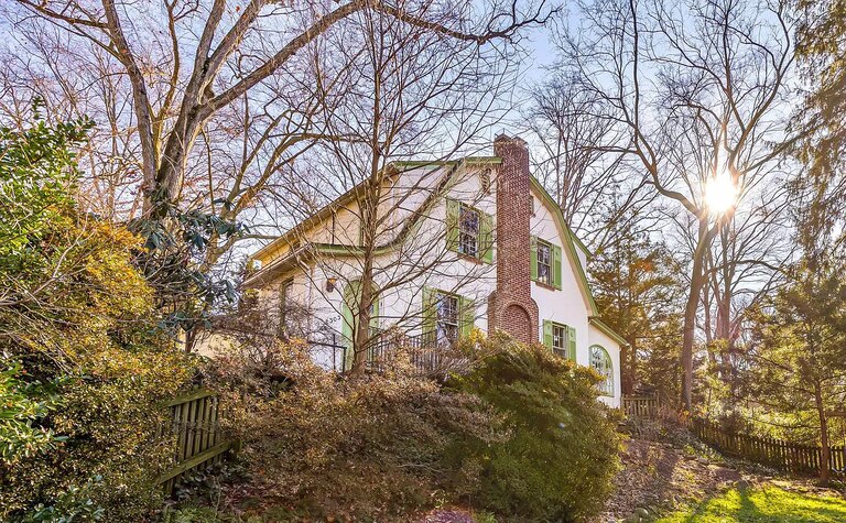 A white house with green shutters and a brick chimney sits on a small hill surrounded by landscaped shrubbery and trees.