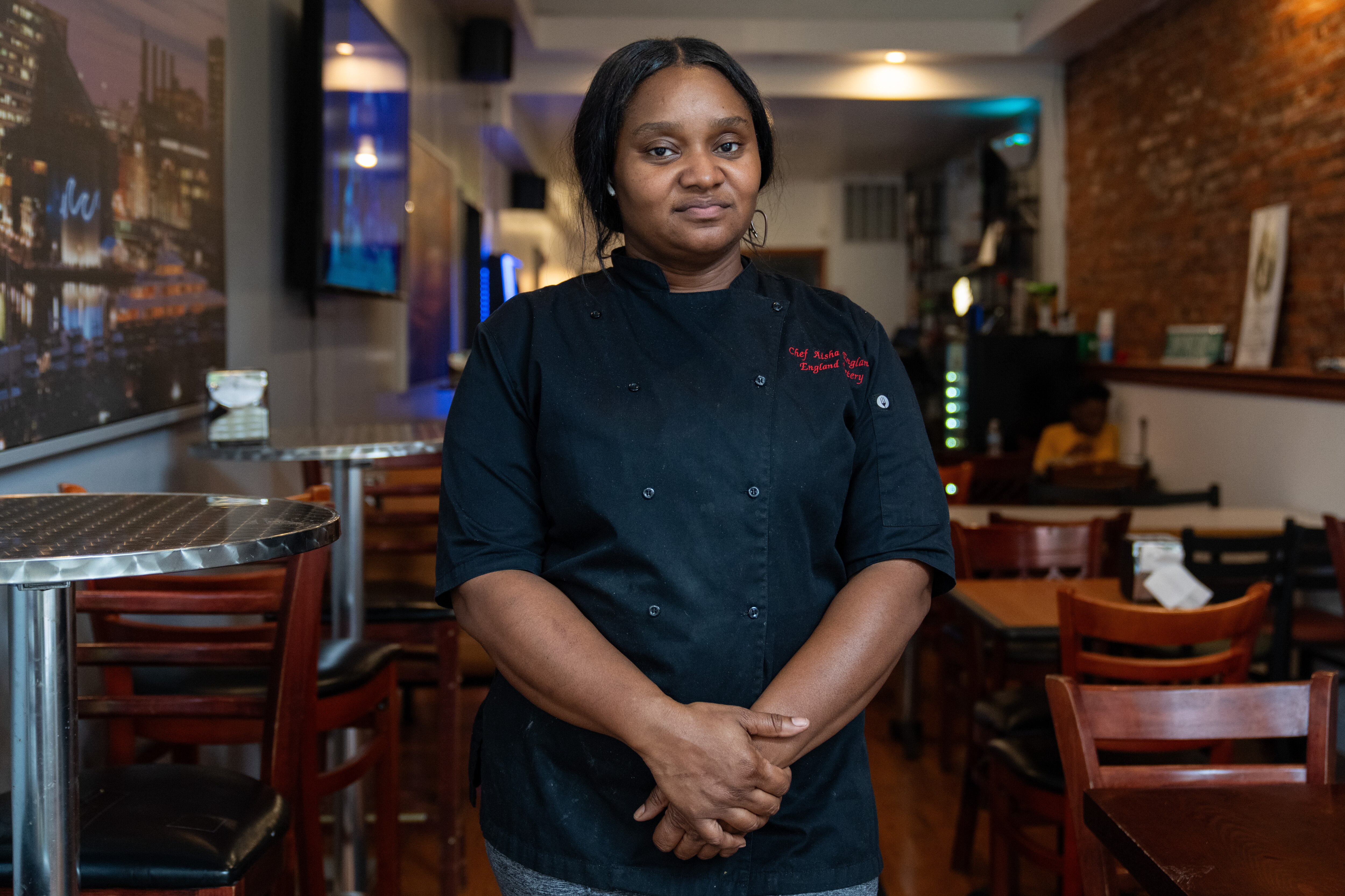 Aisha England poses for a portrait standing in the middle of the restuarant dining area with her hands clasped in front of her.