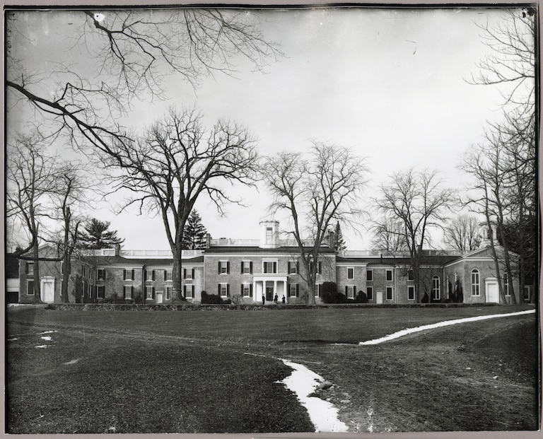 A modern photographic print made from a glass plate negative depicting the exterior of Doughoregan Manor located in Ellicott City, Maryland. Snow is visible on the ground and four figures stand on the steps of the front entrance.