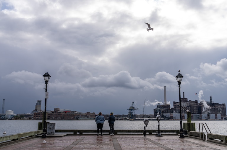 A pair look out at the Patapsco River from Broadway Pier in Fells Point. Dark gray clouds are closest to the horizon, and sunlight breaks through the top of the clouds as a seagull flies by.