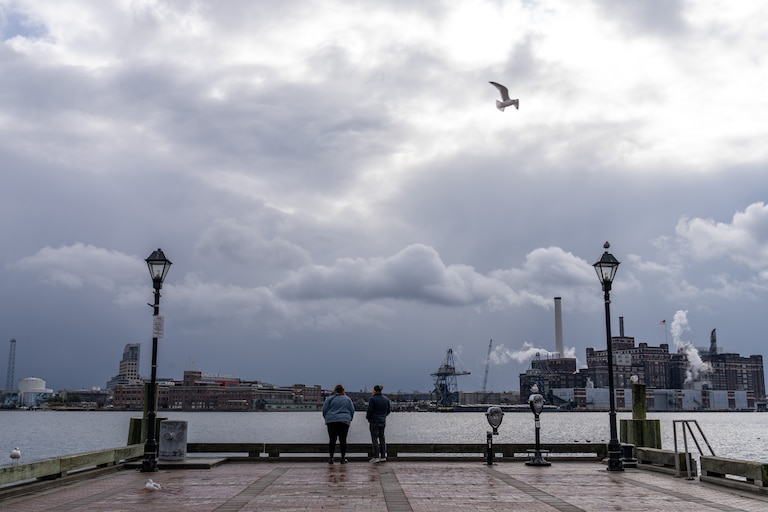 A pair look out at the Patapsco River from Broadway Pier in Fells Point. Dark gray clouds are closest to the horizon, and sunlight breaks through the top of the clouds as a seagull flies by.
