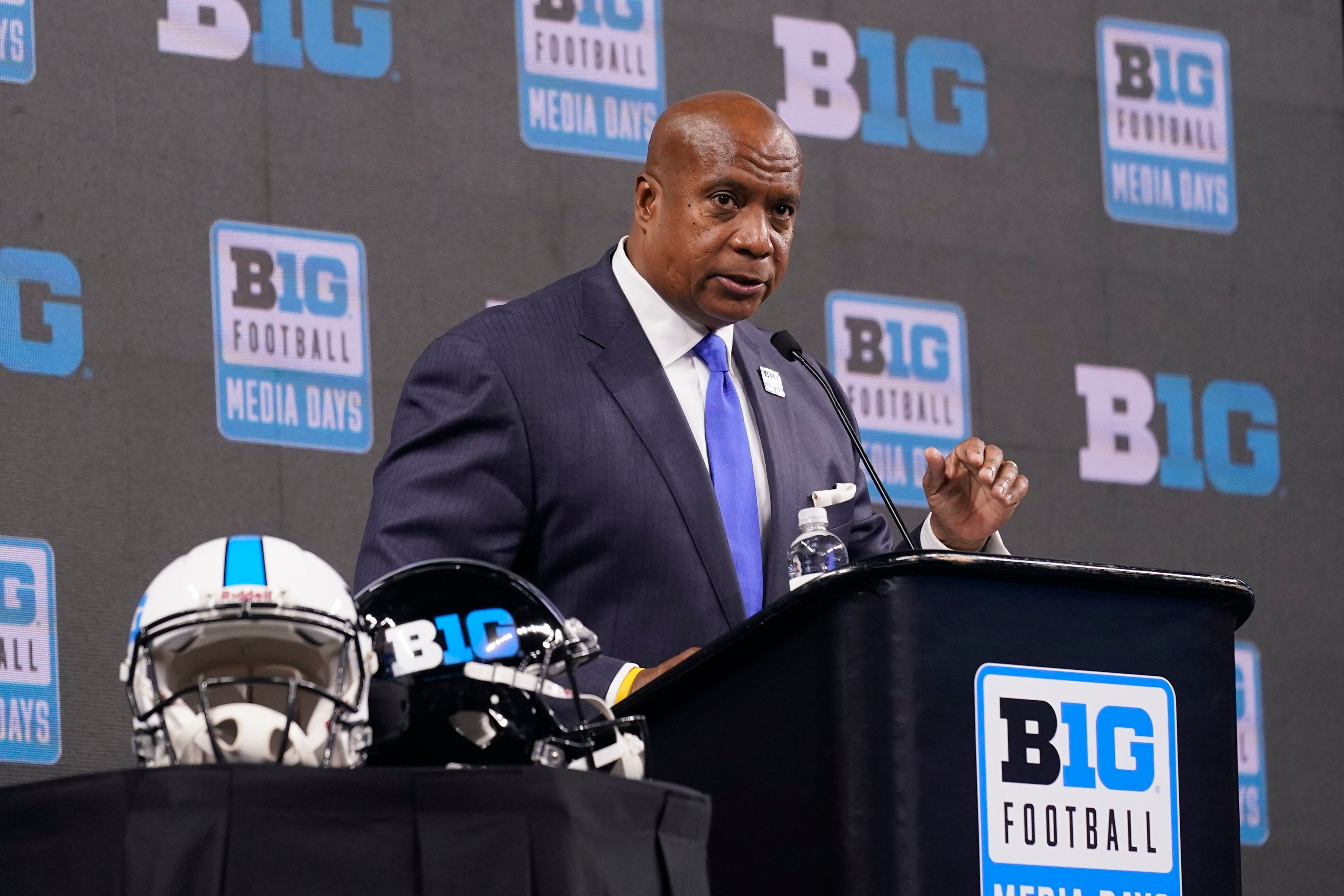 A man in a suit reads from a podium next to two football helmets.