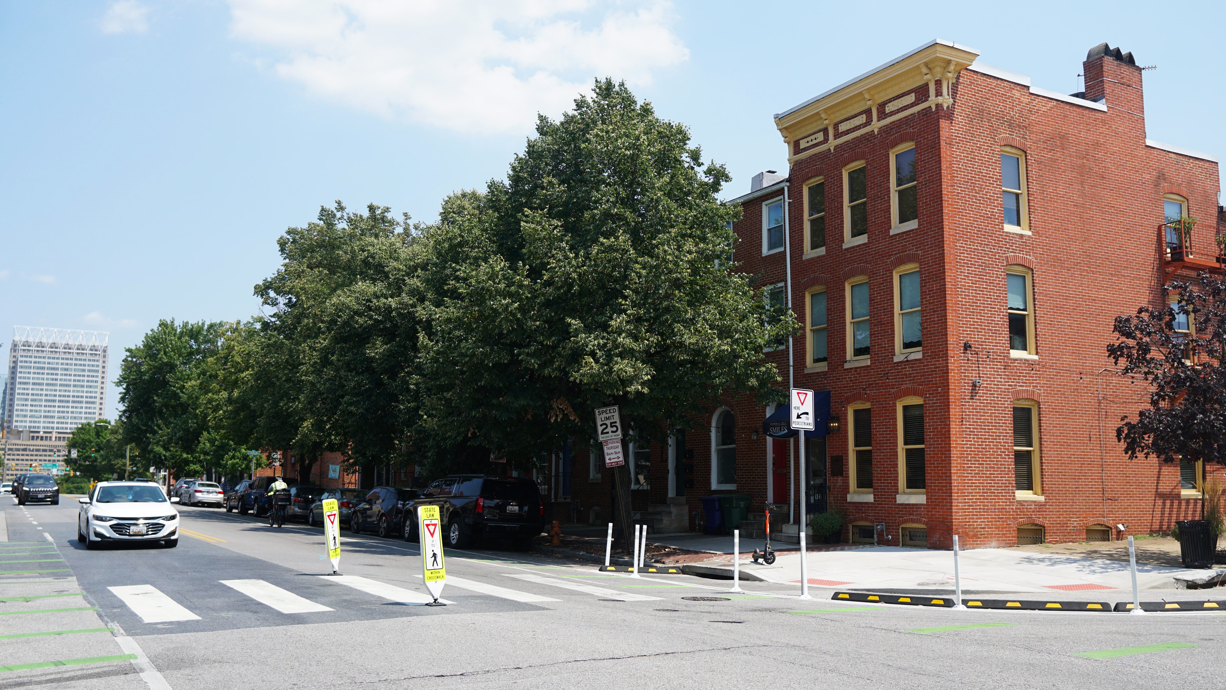 A white car is approaching a city crosswalk on a nice summer day. The crosswalk is in front of a brown, brick building.
