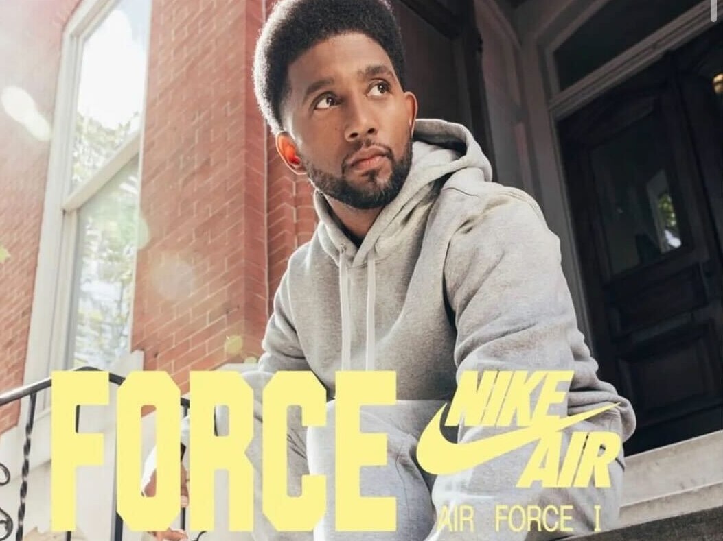 Mayor Brandon Scott sitting on steps in a gray track suit tying the laces on a pair of white Air Force 1 sneakers with a green Nike logo.