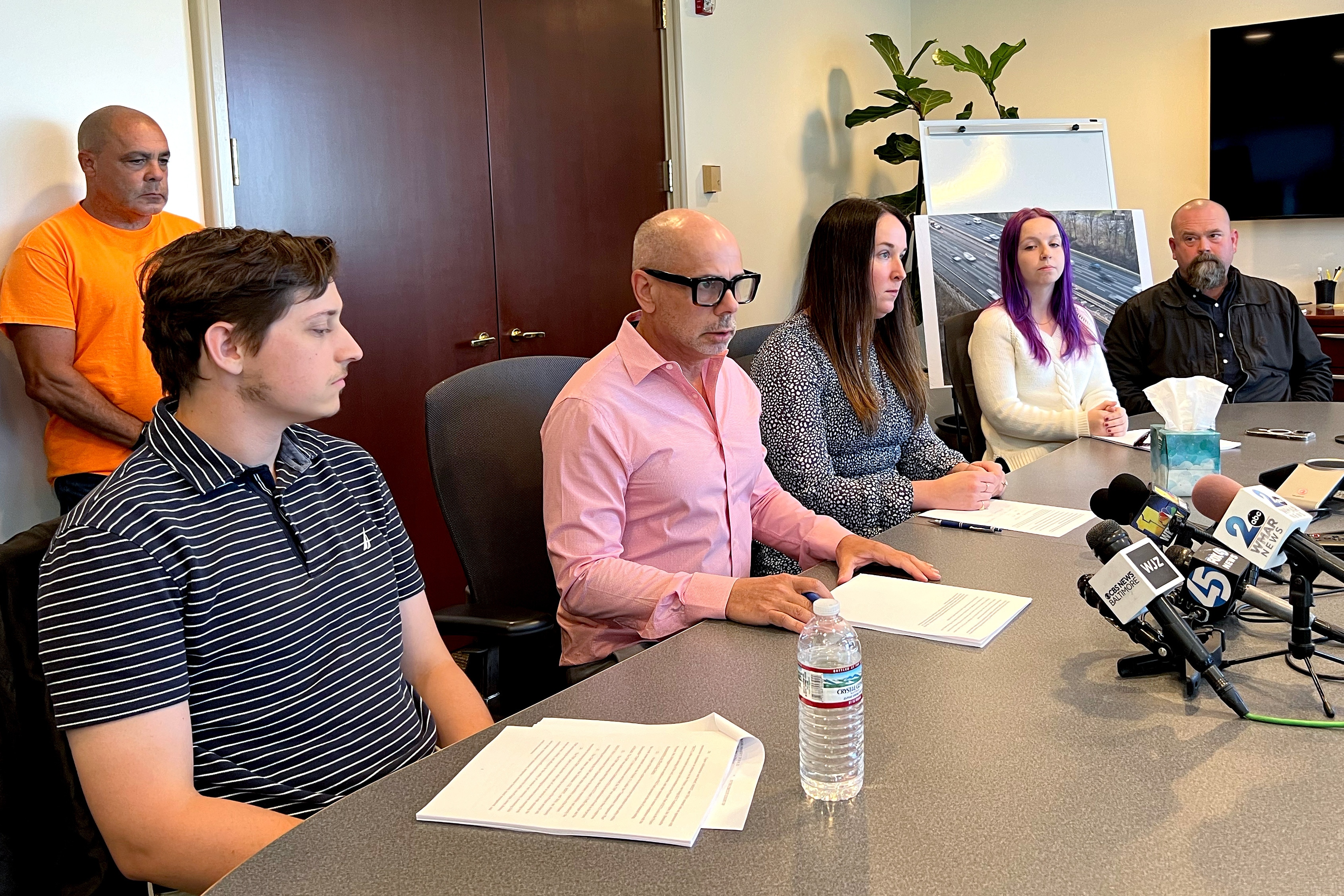 From left: Jim DiMaggio (standing, in orange shirt), Sybil DiMaggio's brother; Dylan DiMaggio, Sybil DiMaggio’s son (seated); Michael Belsky, a partner at Schlachman, Belsky, Weiner & Davey P.A. in Baltimore; Catherine Dickinson, a partner at Schlachman, Belsky, Weiner & Davey P.A.; Nora DiMaggio, Sybil DiMaggio’s daughter, and George Durm III, Sybil DiMaggio’s husband. They held a news conference about the filing of a lawsuit in Baltimore County Circuit Court in a crash in 2023 on Interstate 695 in Baltimore County that killed six construction workers.