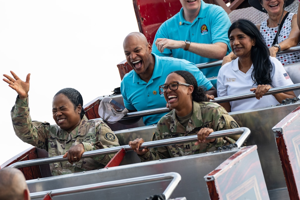 Gov. Wes Moore smiles broadly as he rides a swinging pirate ship attraction. He sits next to Aruna Miller and behind two female Army soldiers.