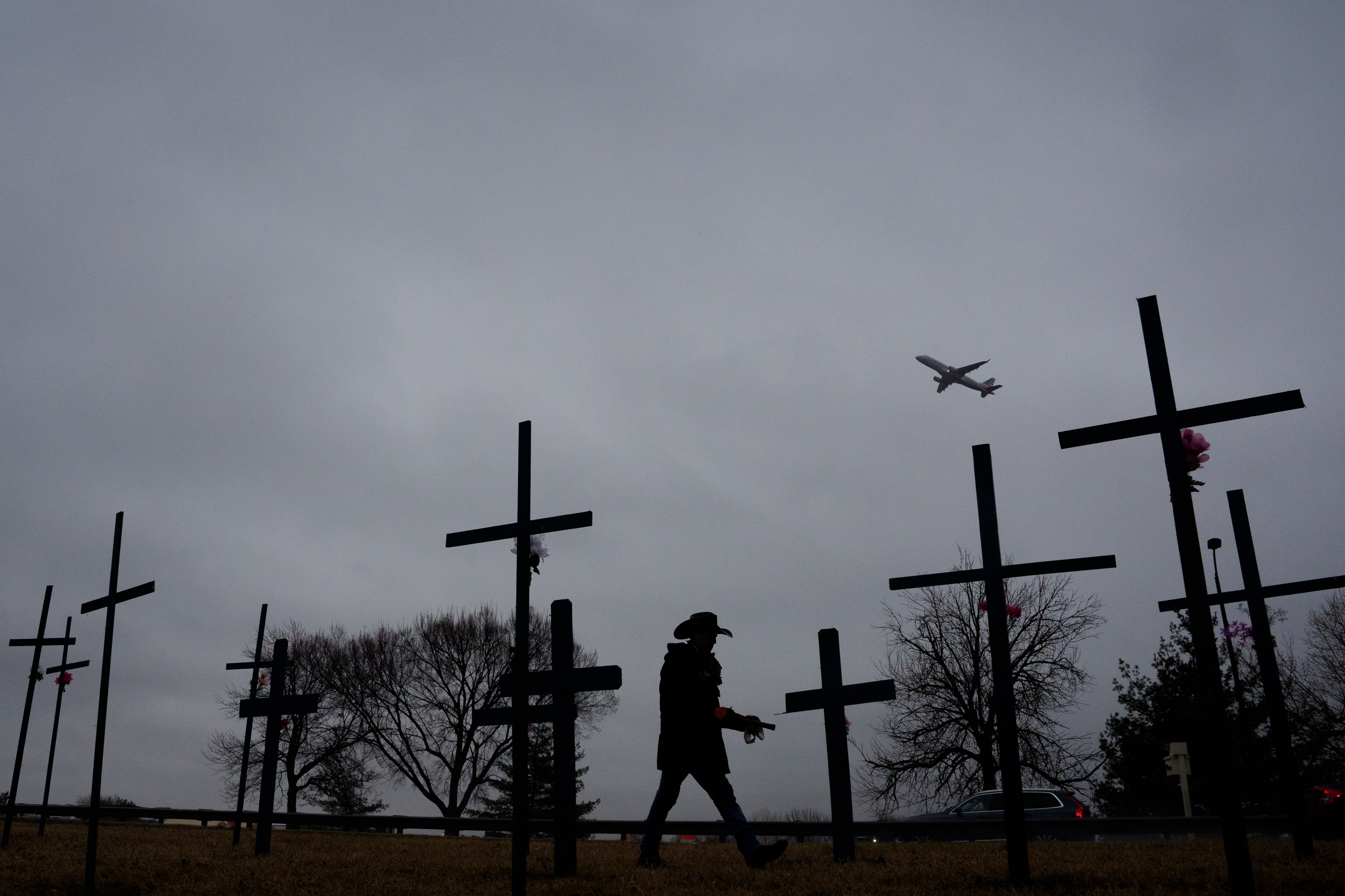 ARLINGTON, VIRGINIA - JANUARY 31: An airplane takes off from Reagan National Airport as Roberto Marquez from Dallas, Texas, puts up crosses as part of a memorial for the victims of the midair collision between an American Airlines plane and a military helicopter earlier this week in the Potomac River, January 31, 2025 in Arlington, Virginia. Flights have resumed in and out of Reagan National Airport after an American Airlines flight from Wichita, Kansas collided midair with a military Black Hawk helicopter while on approach to the airport. According to reports, there were no survivors among the 67 people onboard both aircraft.