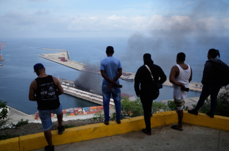 Men watch smoke rising from a dock after explosions were heard at La Guaira port, Venezuela, Saturday, Jan. 3, 2026. (AP Photo/Matias Delacroix)