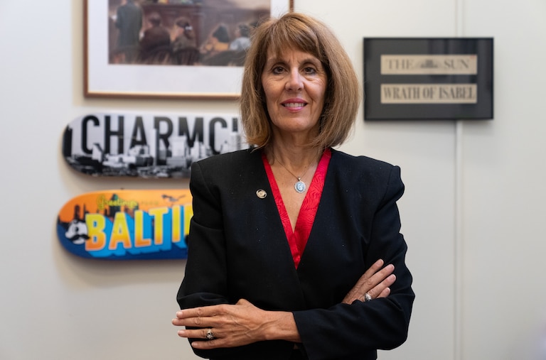 Isabel Cummings poses for a portrait with her arms crossed in her office. Clockwise, the original courtroom sketch of the Jacqueline McLean case she worked on, a Baltimore Sun clipping reading "Wrath of Isabel," a blue and orange "Baltimore" skateboard and a black and white "Charm City" skateboard hang behind her.