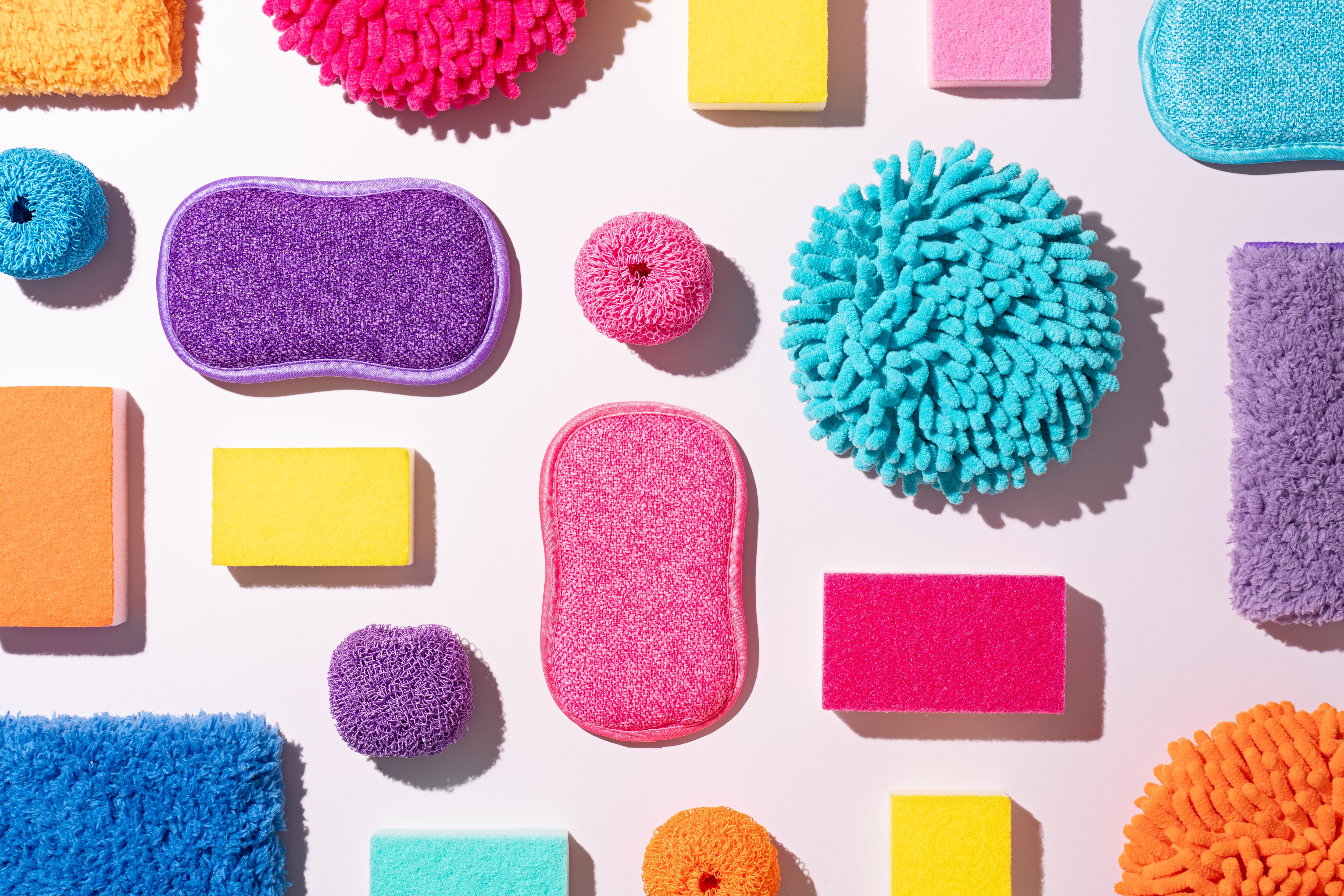 A Collection of Multi Colored Cleaning Supplies Collection Includes Chenille Microfiber Cleaning Cloth, Rag, Scouring Pad, Cleaning Sponge, Dish Cloth and Dish Towel on Pink Background Directly above View.