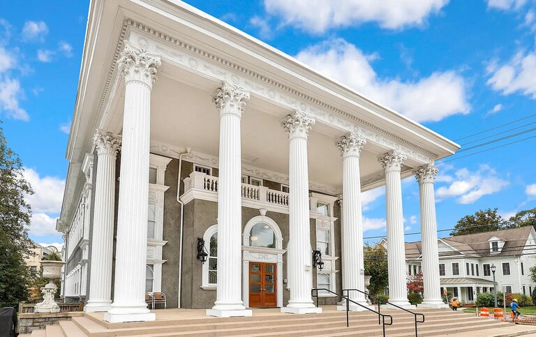 A street-level view of an old gray building with white trim, a chestnut colored door and massive white columns appears against a blue sky with clouds.