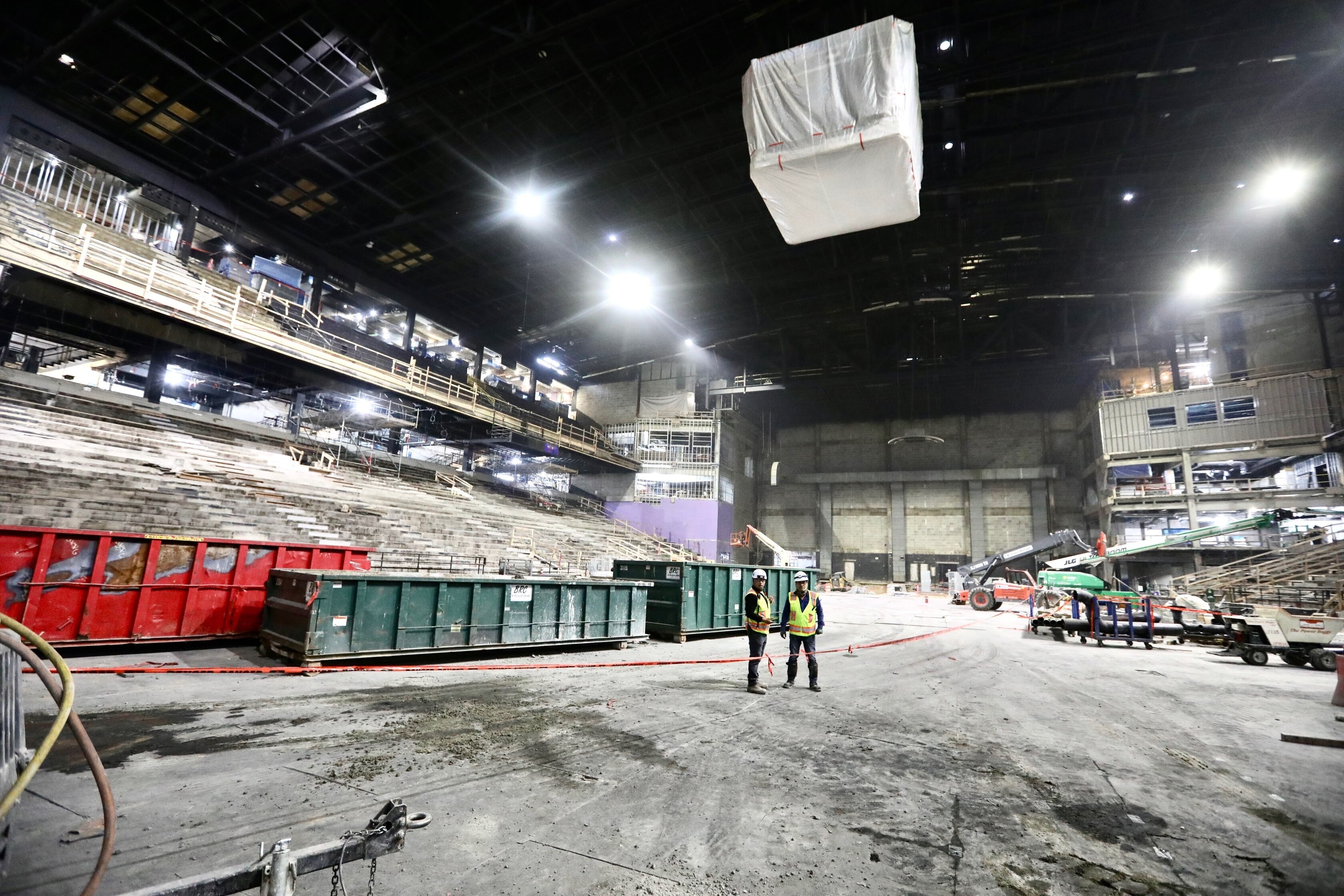 Two workers wearing hard hats and vests stand amid metal trash receptacles inside CFG Bank Arena.