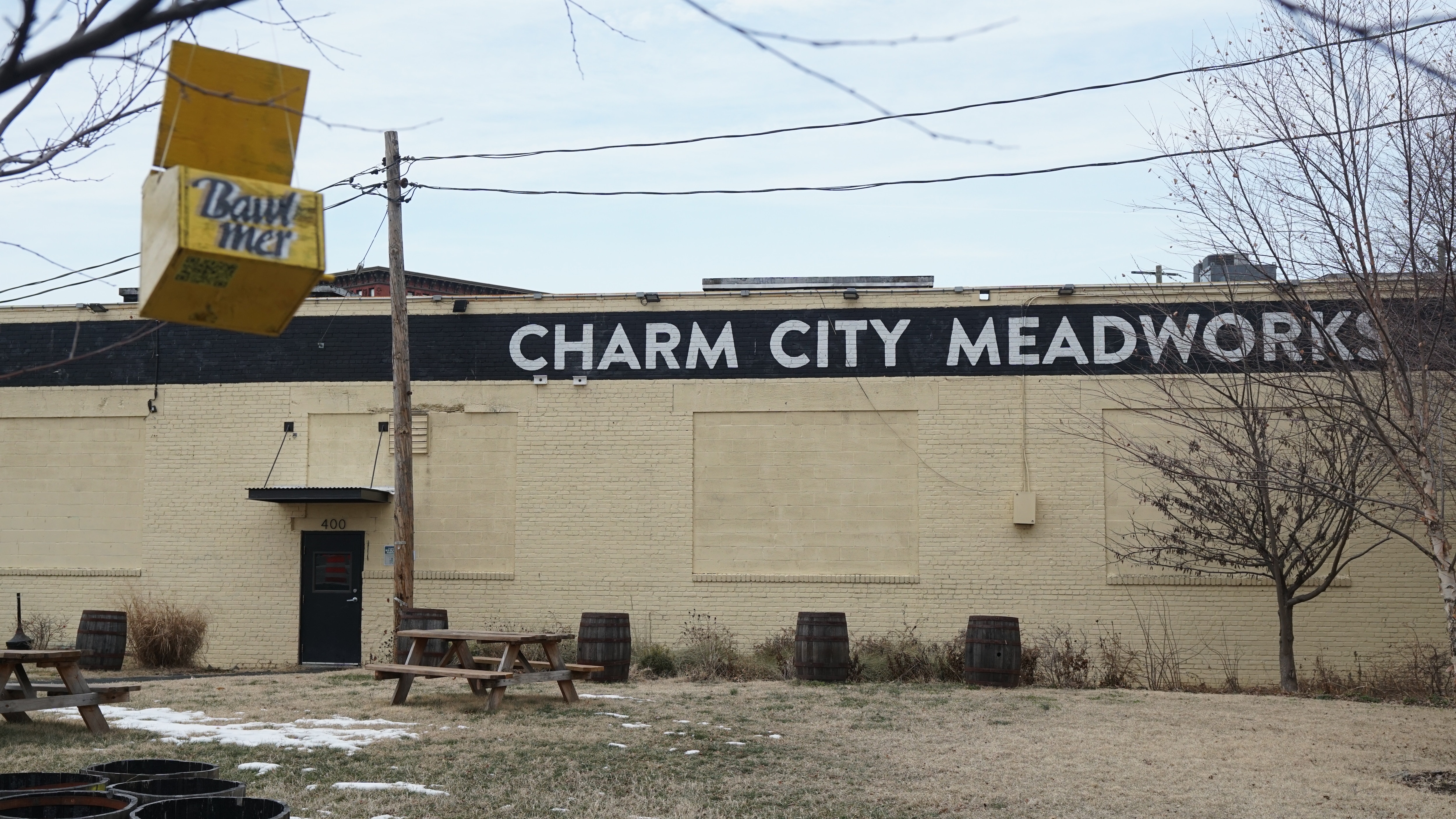An industrial-looking building that reads "CHARM CITY MEADWORKS" at the top is in the background and a small yellow bird feeder that reads "BAWLMER" swings from a tree branch in the foreground.