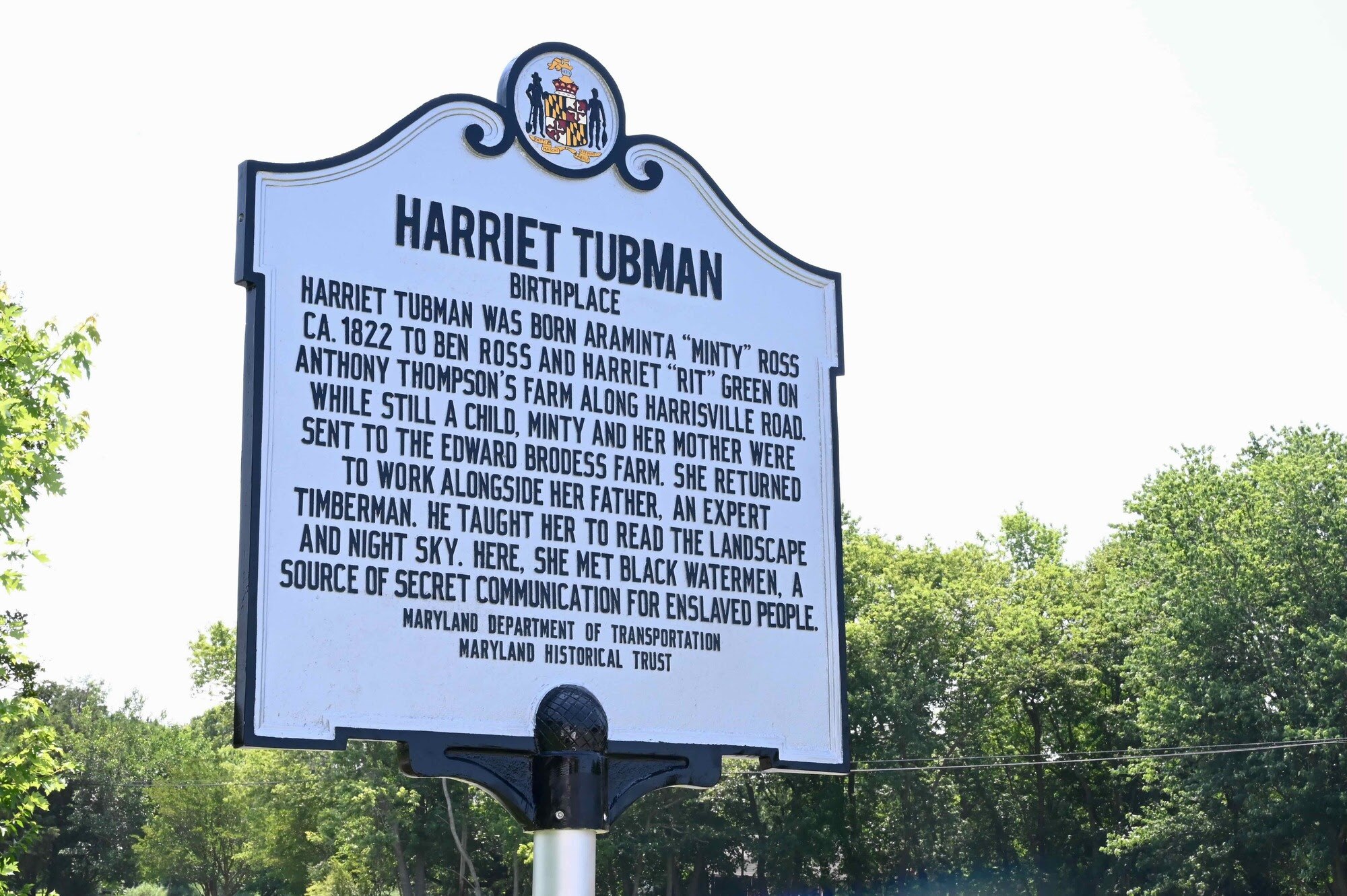 A roadside sign commemorating Harriet Tubman's birthplace on the Eastern Shore. The sign reads: arriet Tubman was born Araminta "Minty" Ross CA. 1822 to Ben Ross and Harriet "Rit" Green on Anthony Thompson's farm along Harrisville Road. While still a child, Minty and her mother were sent to the Edward Brodess farm. She returned to work alongside her father, an expert timberman. He taught her to read the landscape and night sky. Here, she met black watermen, a source of secret communication for enslaved people.