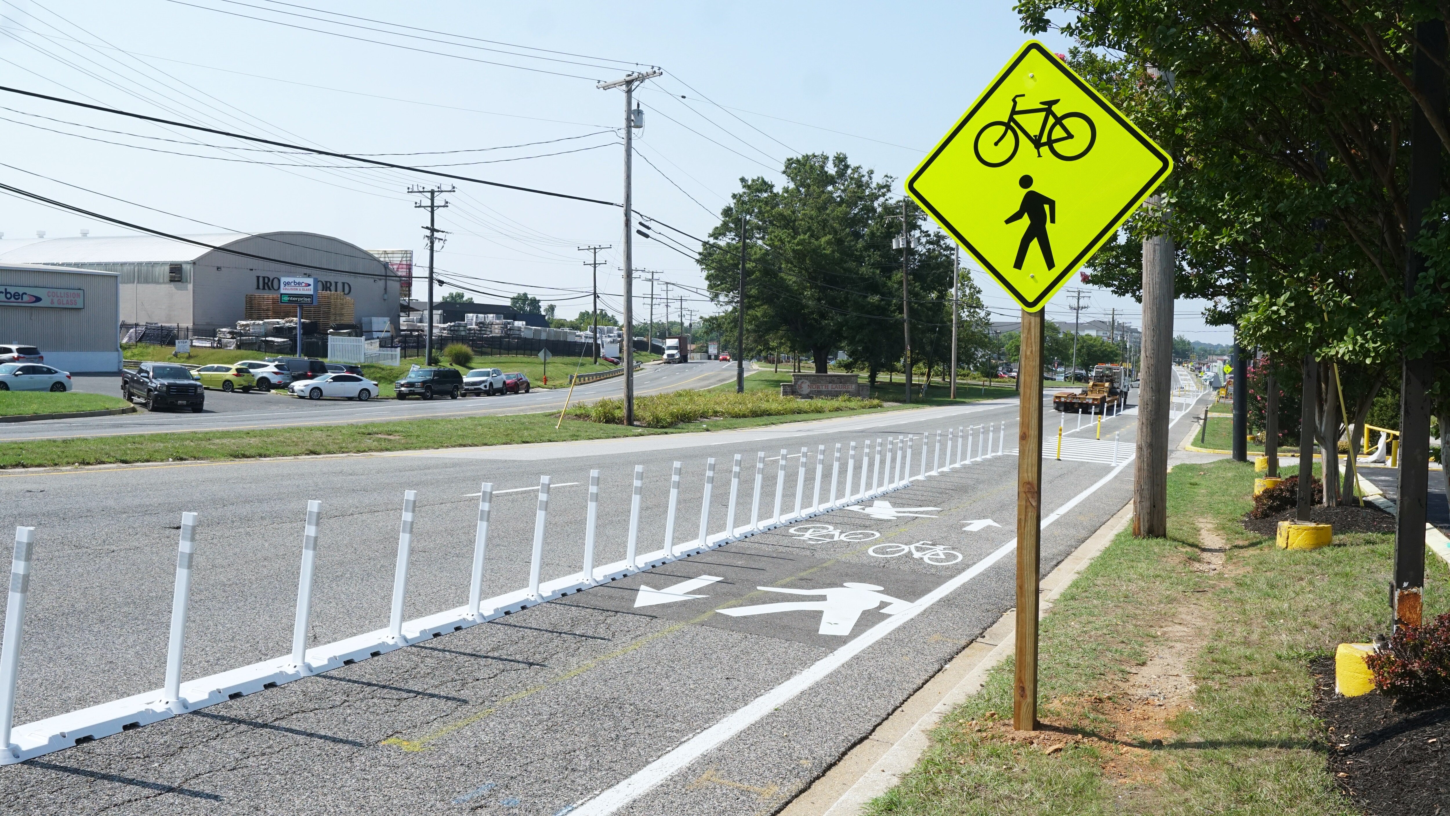 A bike lane is seen sectioned off from the road by white plastic flex posts. A yellow sign telling drivers to pay attention to bikers and pedestrians is on the side of the road.