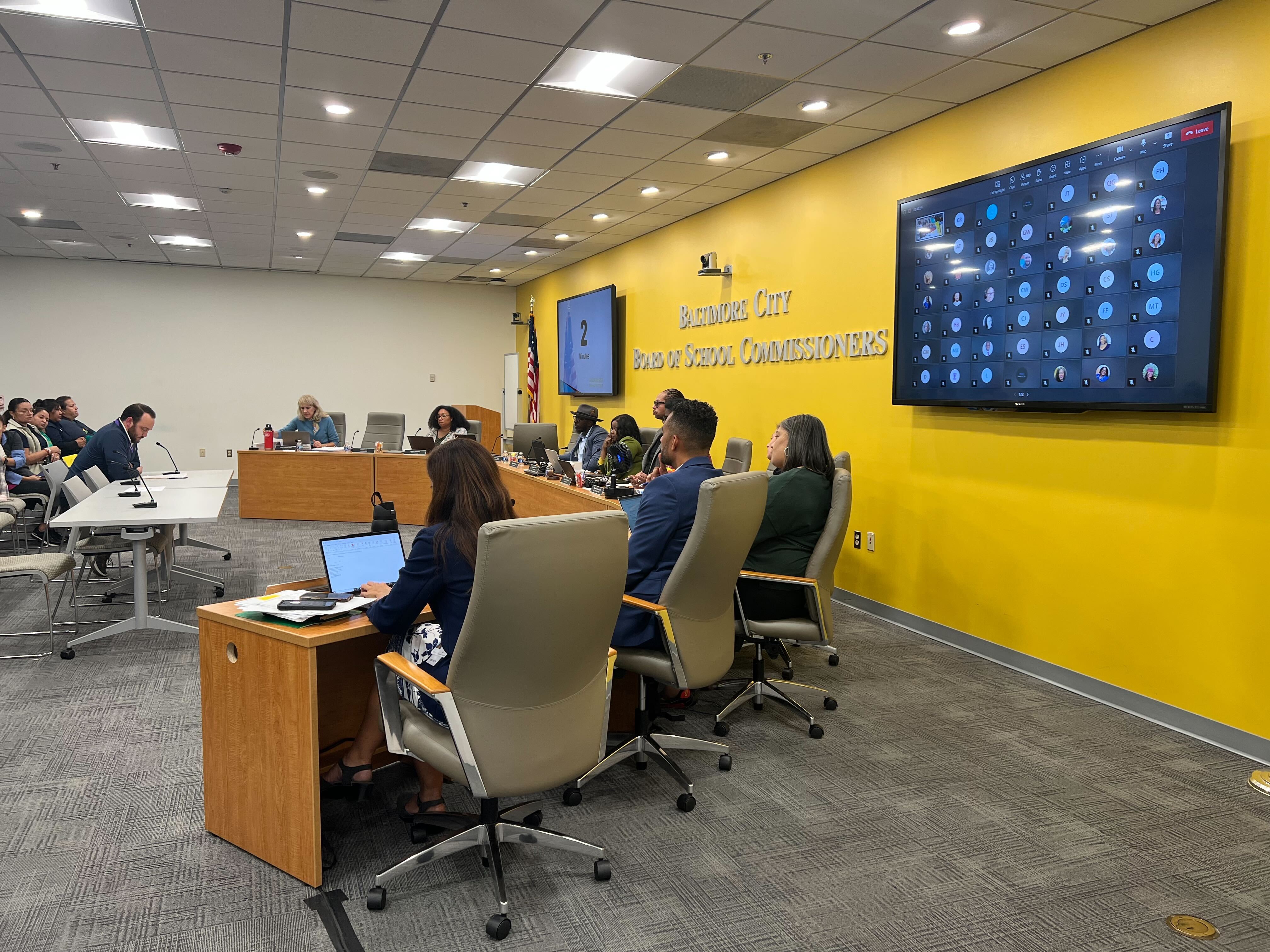 A group of people sit in high-backed chairs behind a curved desk. A man is in front of them sitting at a table and speaking into a microphone.