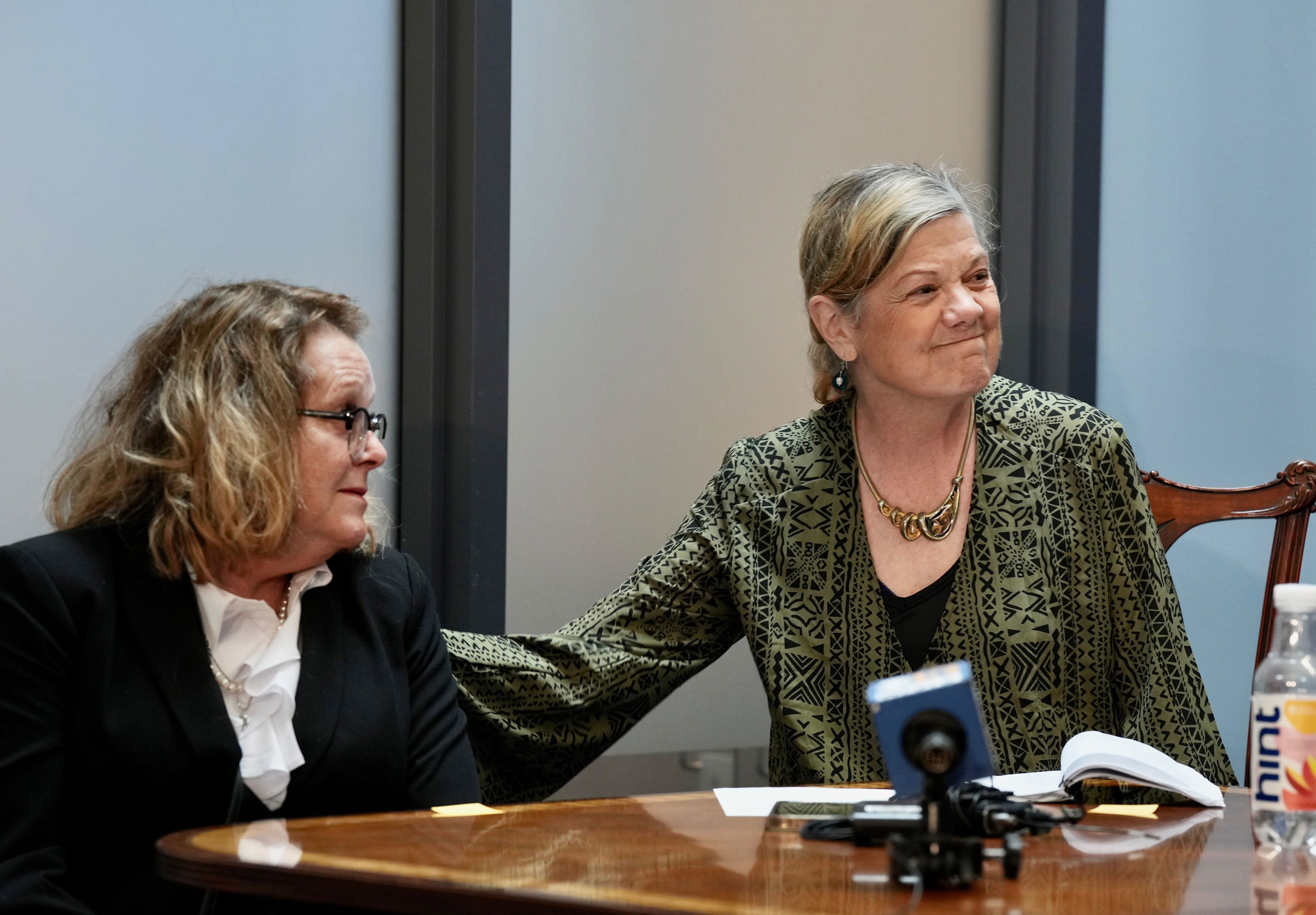 A woman wearing a green shirt smiles while putting her right hand on the back of a woman wearing a black blazer and white shirt.