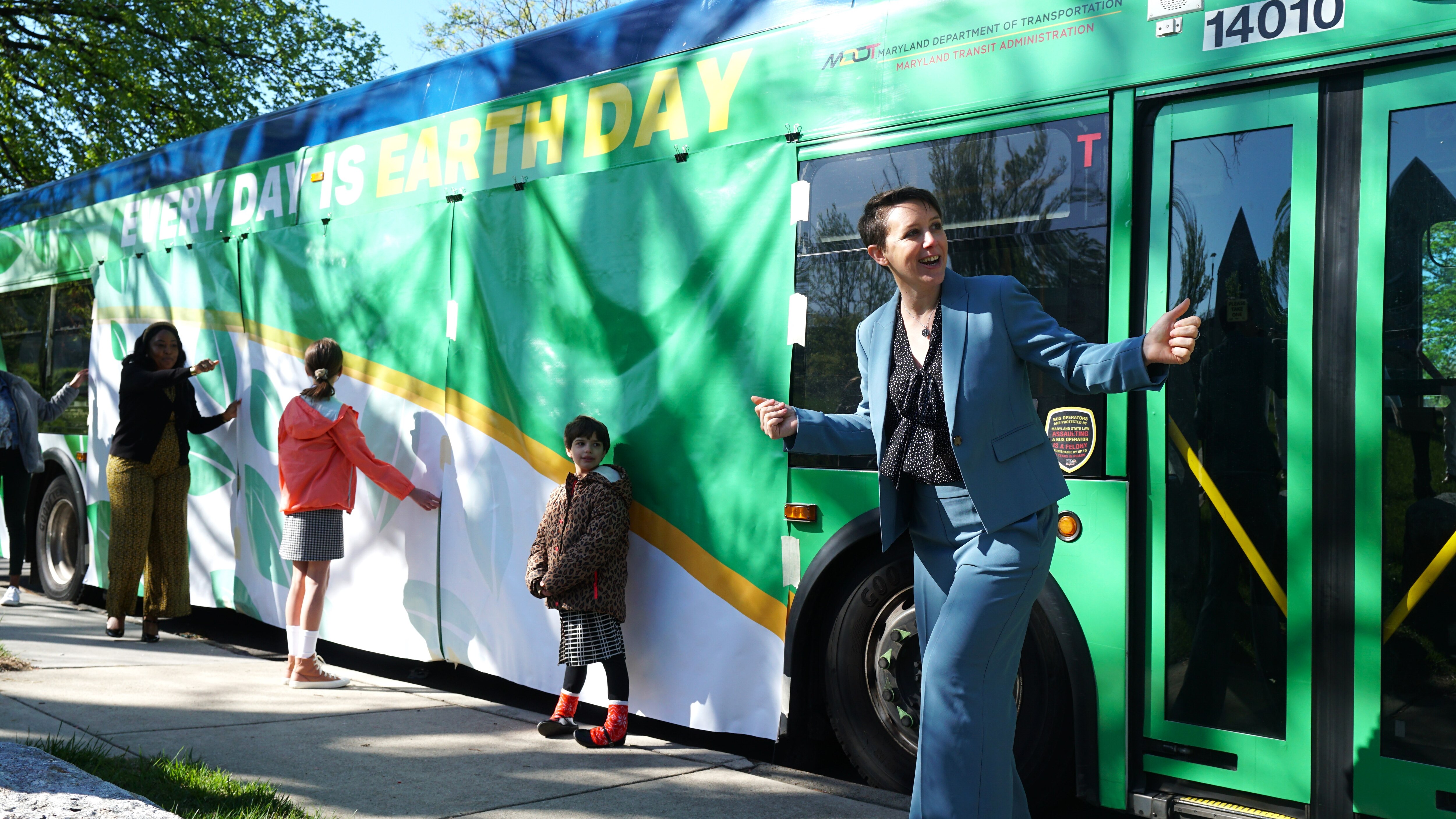 A woman in a blue suit stands in front of a green bus. Two kids stand in front of green and white panels covering the side of the bus.