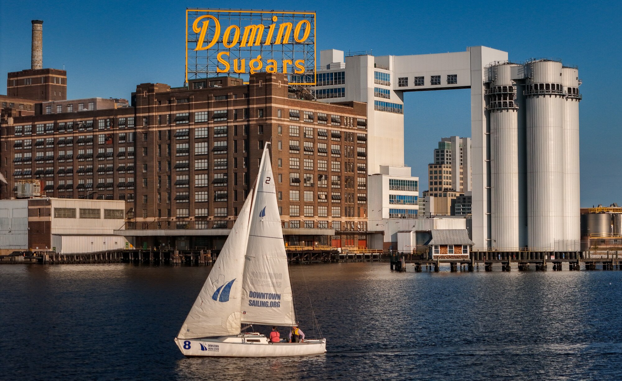 Members of the Downtown Sailing Center sail past the Domino Sugars plant during a late afternoon outing on Baltimore’s Inner Harbor.