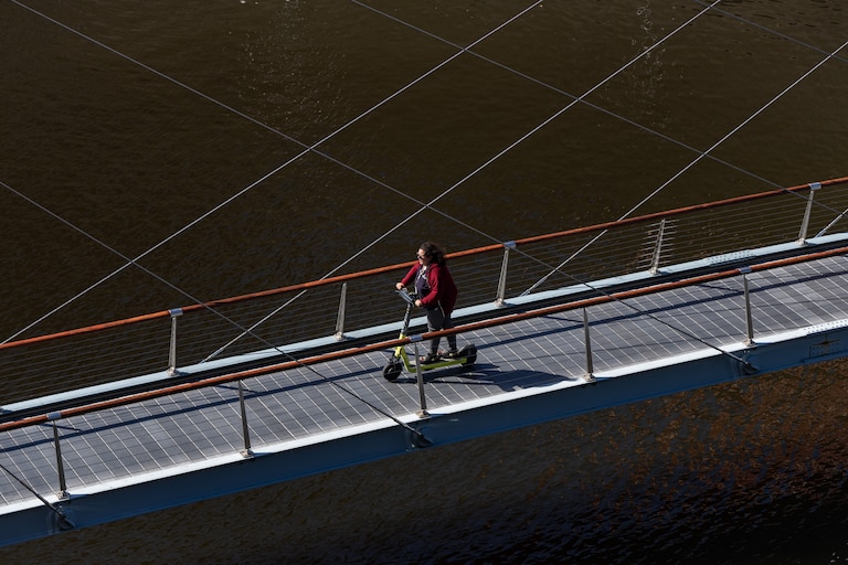 A woman scootering across the Harbor Bridge Walk Path is framed between the cables supporting the bridge.