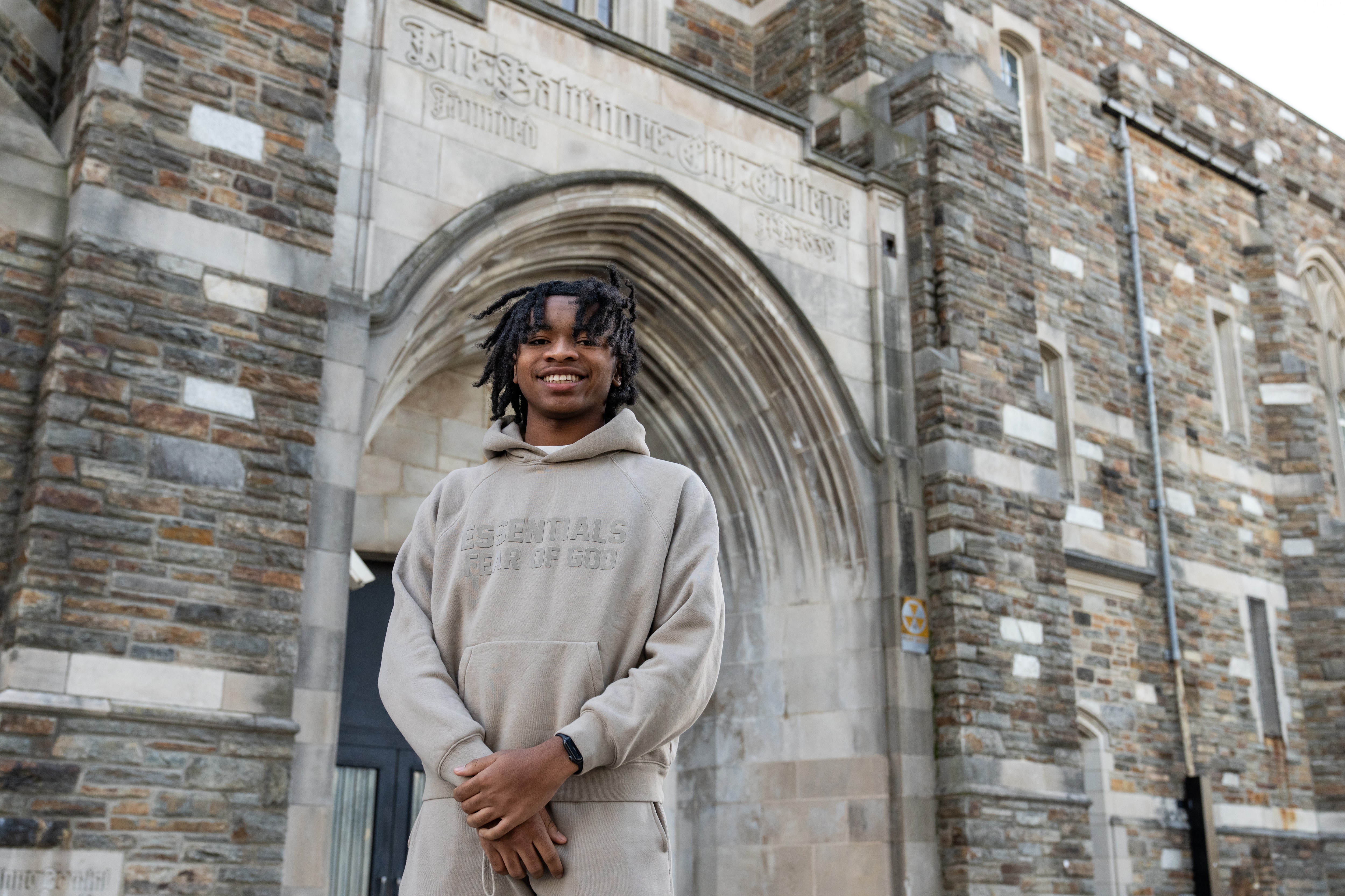 Travis Winstead Jr. smiles for a portrait in front of a stone archway that reads "The Baltimore City College."