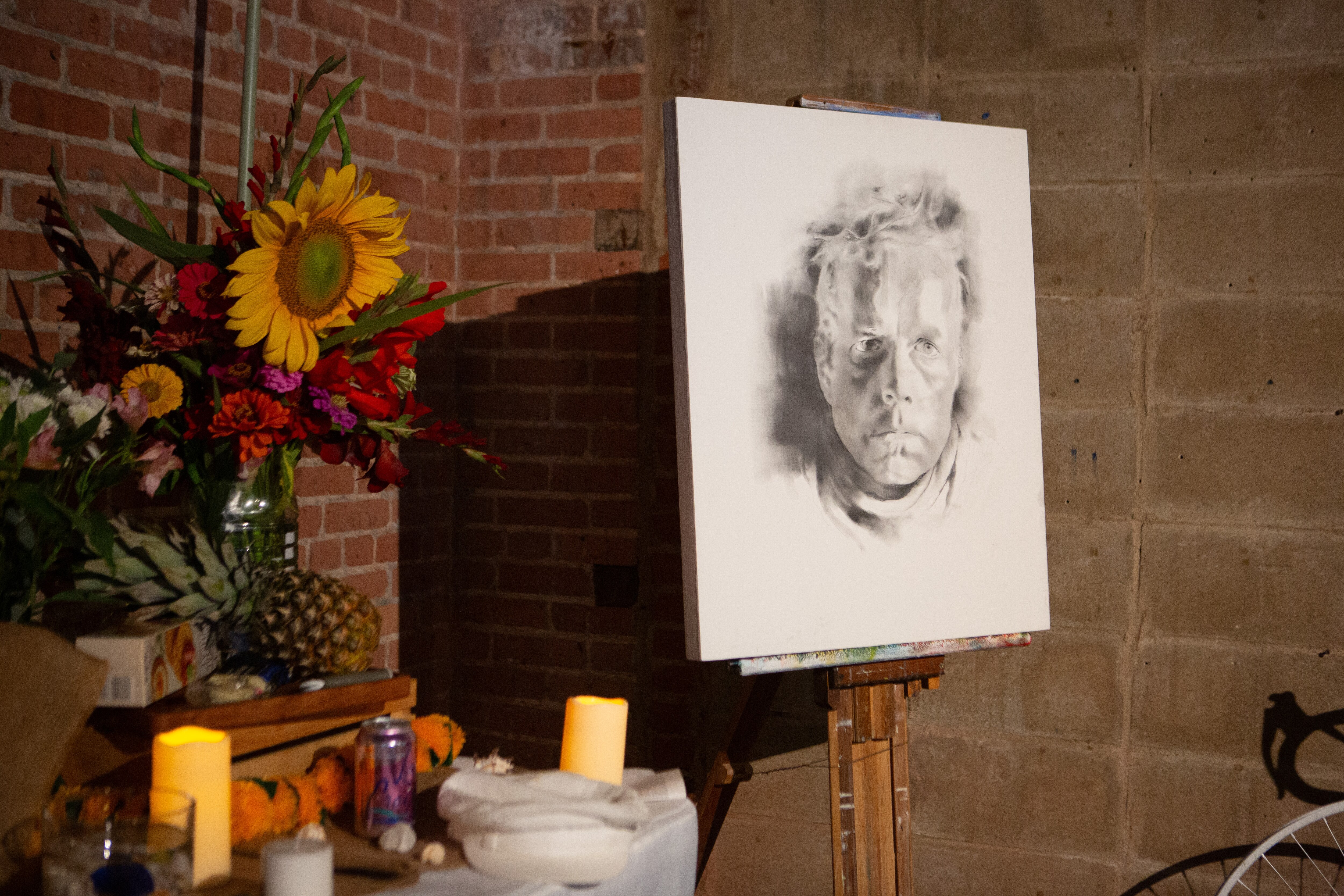 A self portrait of David Herman stands next to a table of mementos from friends, including his favorite snack of cheese and crackers, flowers, and a can of LaCroix sparkling water.